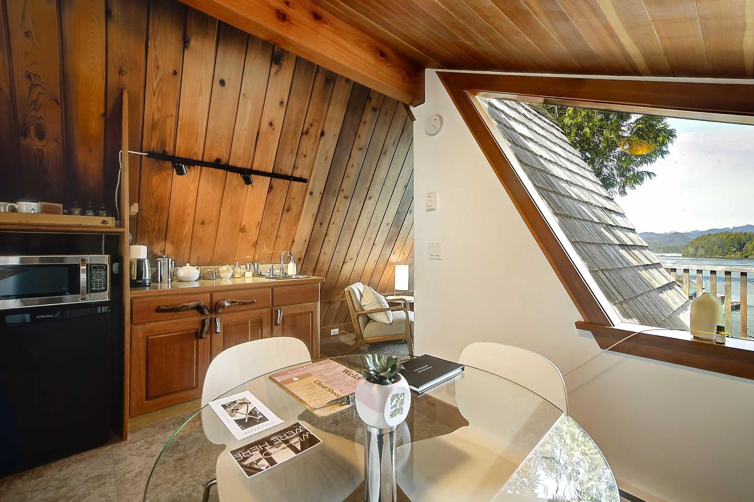 Interior view of a kitchen and dining area with wooden paneling on the ceiling, a large triangular window showing trees and a lake outside, and a glass table with magazines, a plant, and some papers. Kitchen cabinets and appliances are visible on the