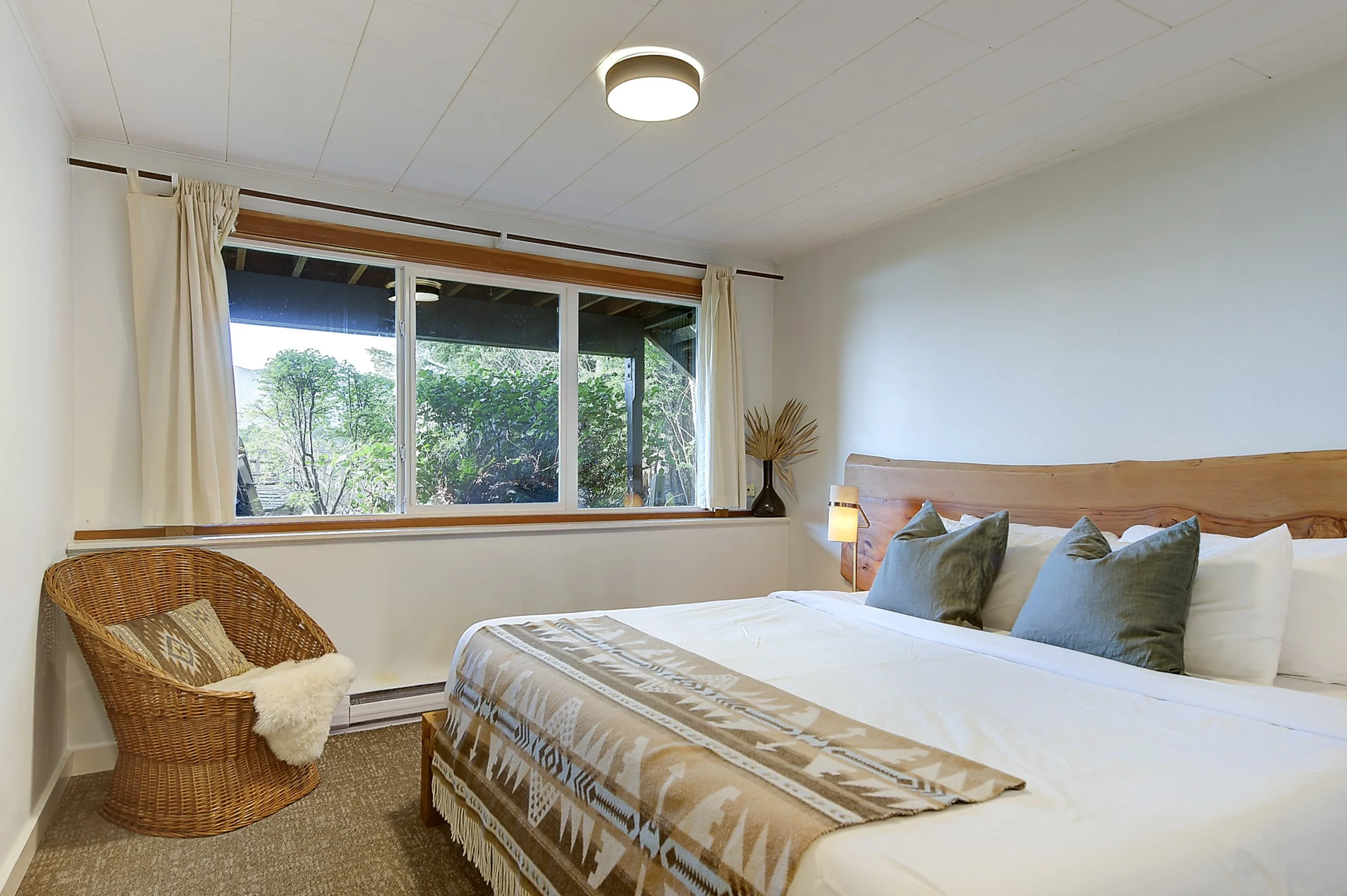 A bedroom with large window, white walls, a wooden headboard, bedside lamp, decorative plants, and woven chair.