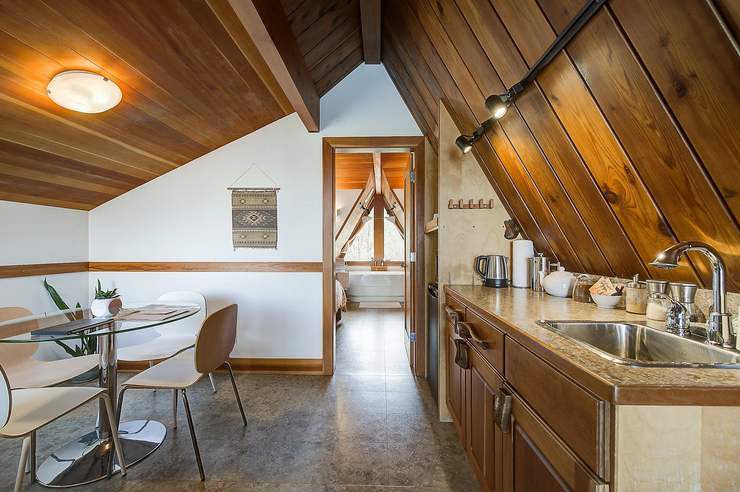 Interior view of an attic-style kitchen with wooden ceiling panels, a small dining table, and a doorway leading to a bathroom with a bathtub.
