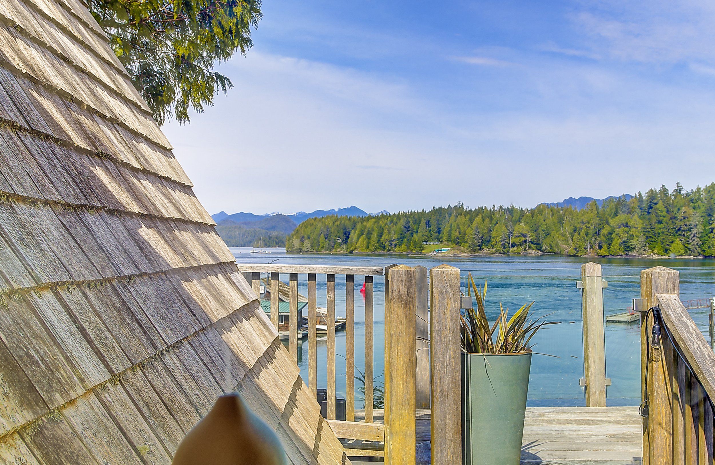 A lakeside scene with wooden shingle roof in the foreground, a wooden fence, and a body of water with trees on the opposite shore. Mountain peaks are visible in the distance under a blue sky with some clouds.
