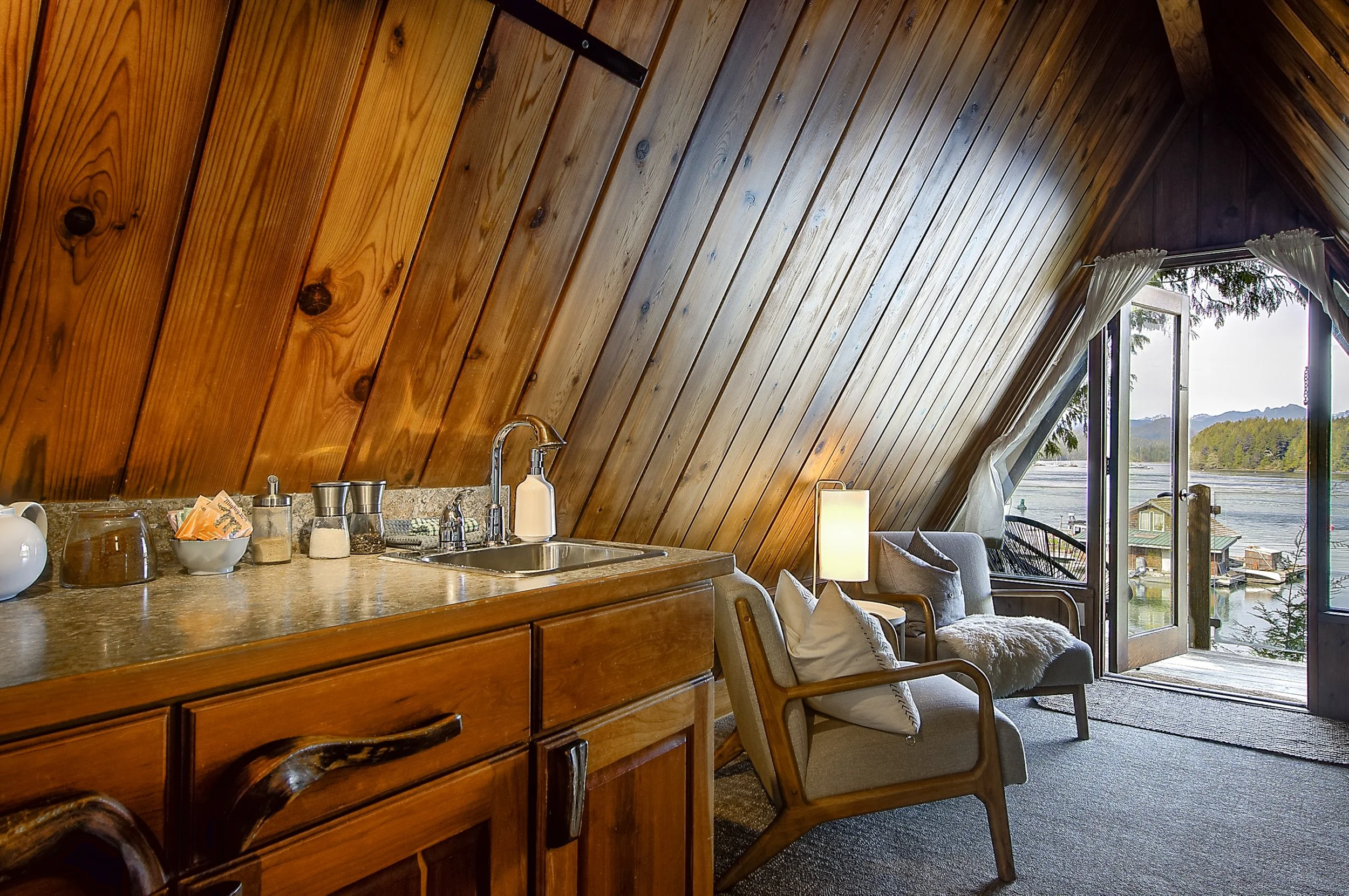 Interior view of a cabin or lake house with a slanted wooden ceiling, kitchen counter with items, and a sitting area with armchair and large window overlooking water and trees.