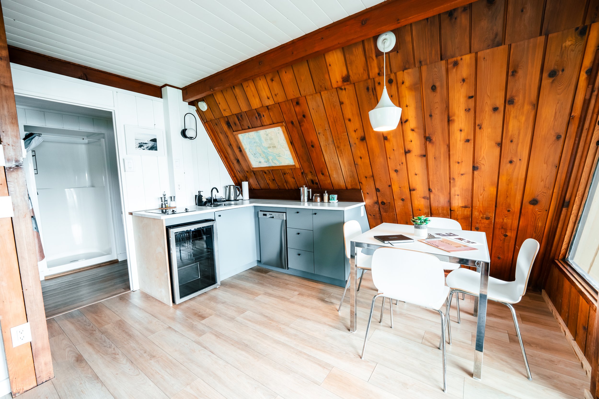 A cozy kitchen and dining area with wood-paneled, vaulted ceiling, white walls, small white table with four white chairs, kitchen counter with appliances, and a window letting in natural light.