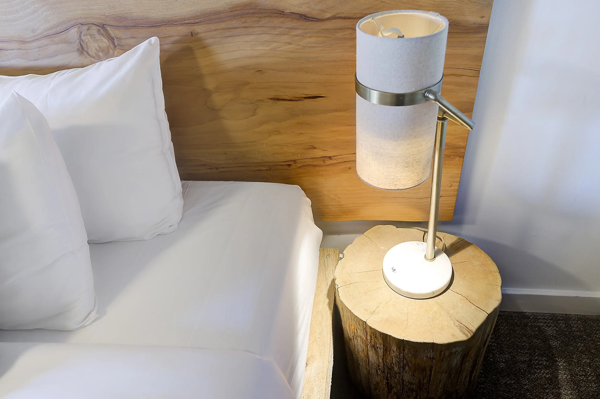 Close up of a bed with white pillows and a wooden headboard. A bedside table made from a cross section of a tree trunk holds a lamp with a cylindrical fabric shade and a metal arm.