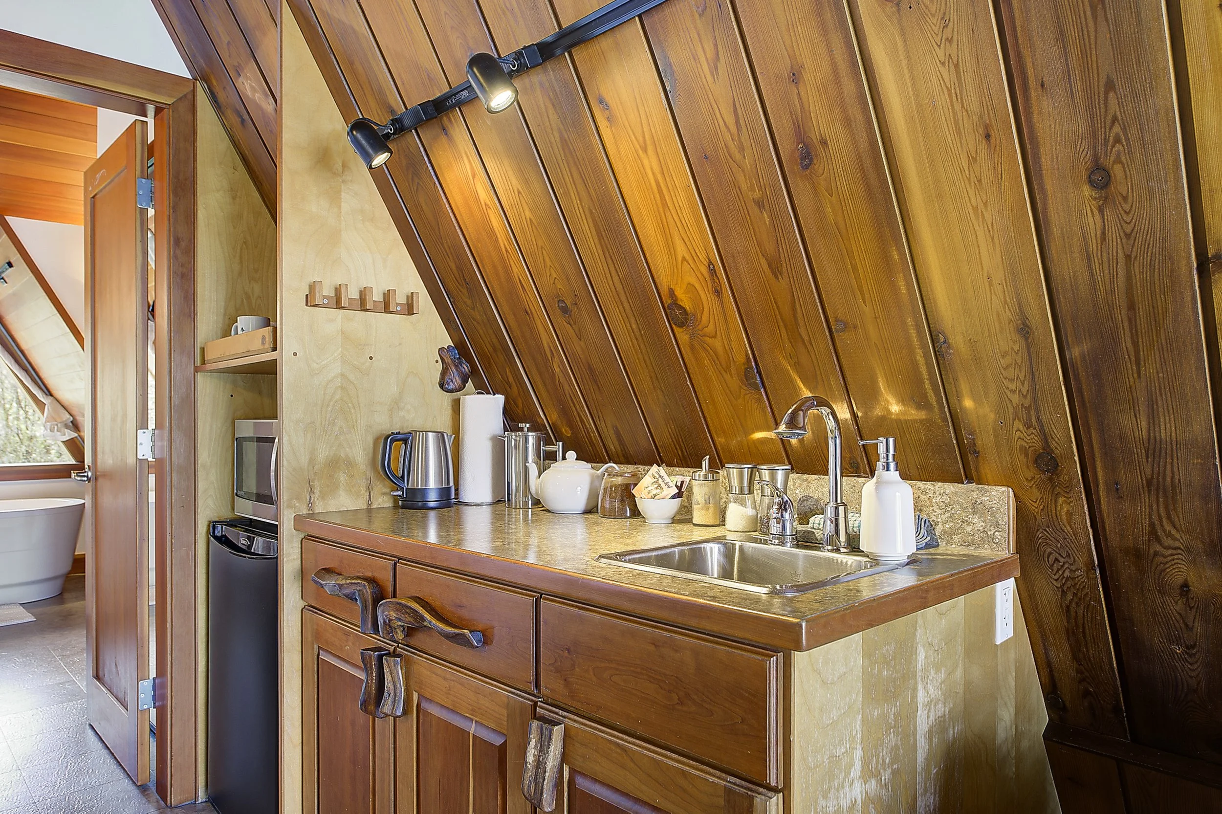 Kitchen area with wood paneling on the ceiling and walls, featuring a stainless steel sink, a soap dispenser, and various kitchen items such as a teapot, cups, and a electric kettle on the countertop. There is a wood shelf with a mug and a small deco