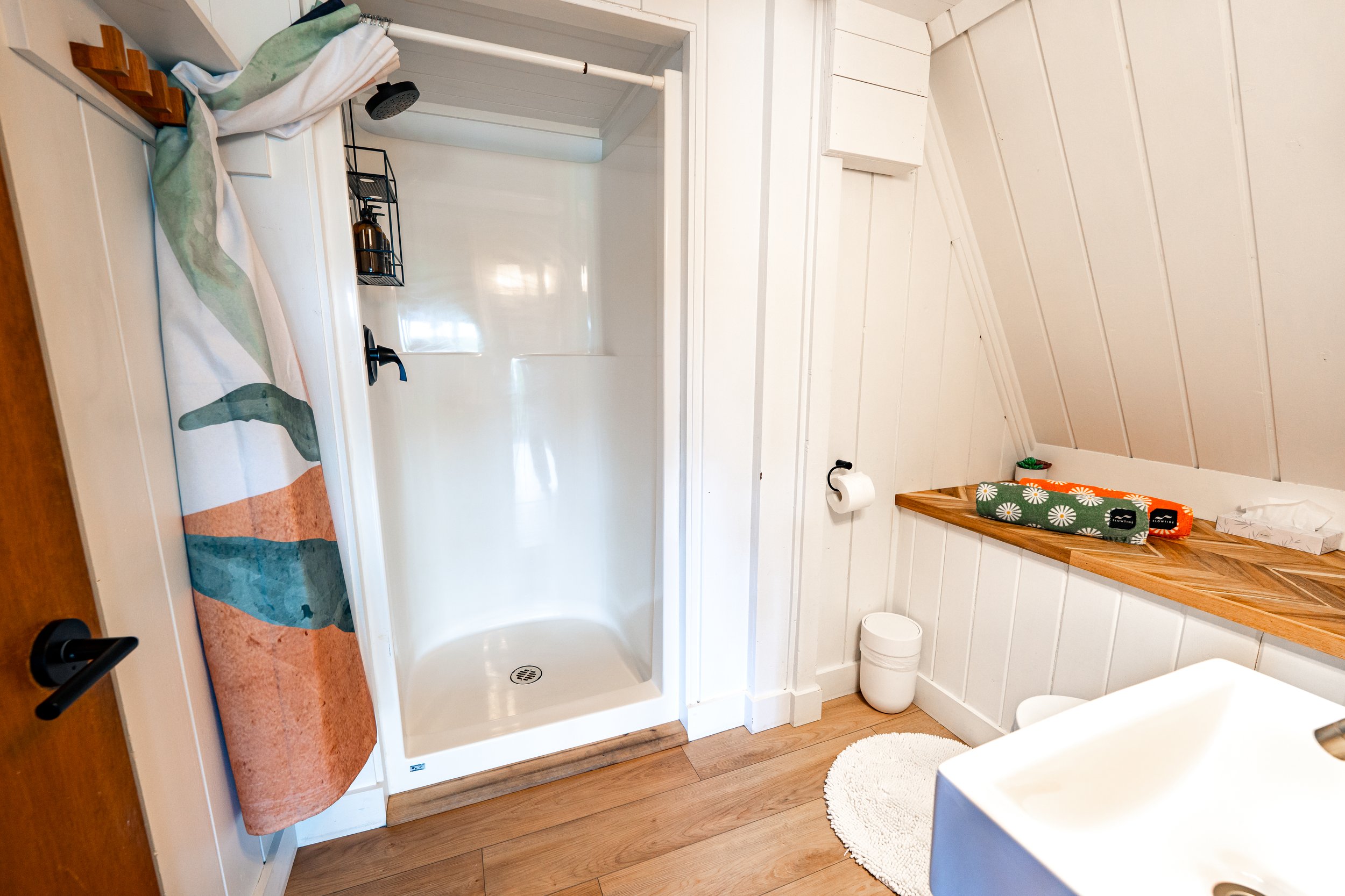 A small bathroom with a shower stall, white beadboard walls, a wooden countertop with a floral towel, and a white round rug on the wooden floor.