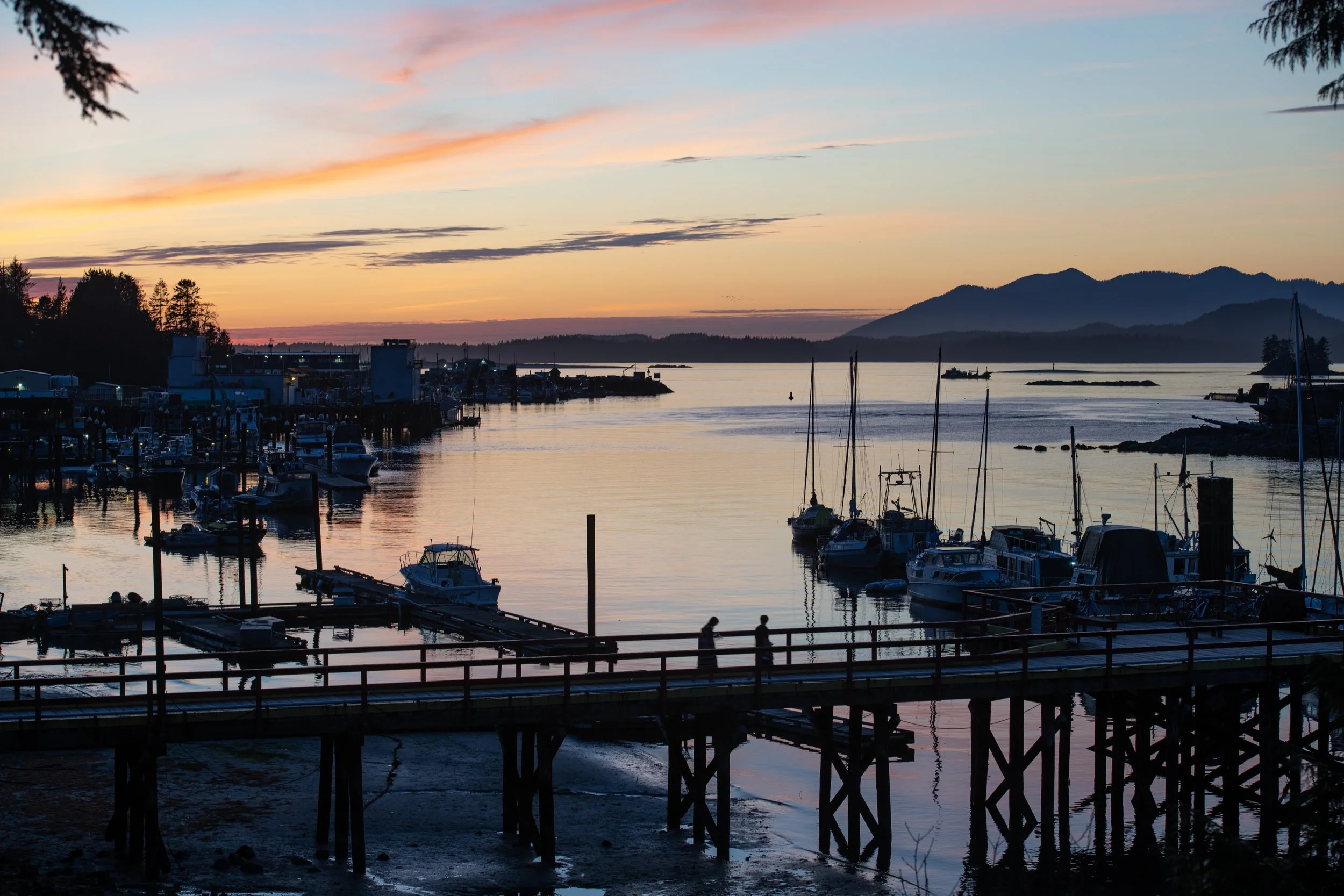 Sunset over a marina with boats docked, mountains in the background, and reflections on the water.