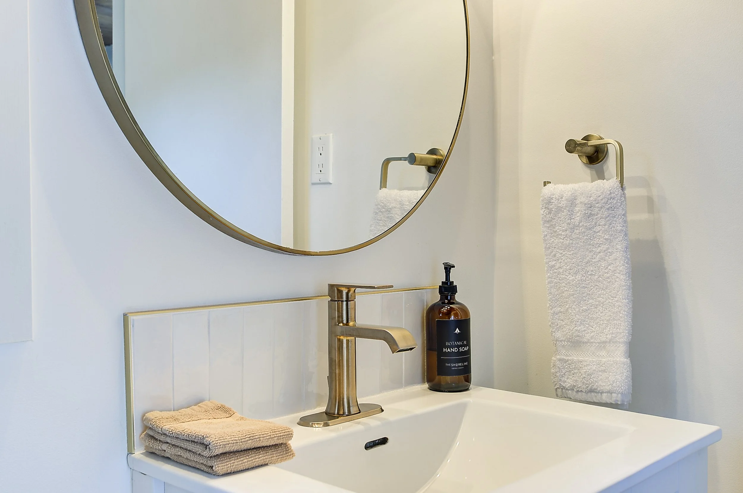 A bathroom sink with a modern gold faucet, a soap dispenser labeled 'Botanical Hand Soap,' a folded beige towel, a mirror above the sink, a towel hanging on a gold towel bar, and a wall outlet near the mirror.