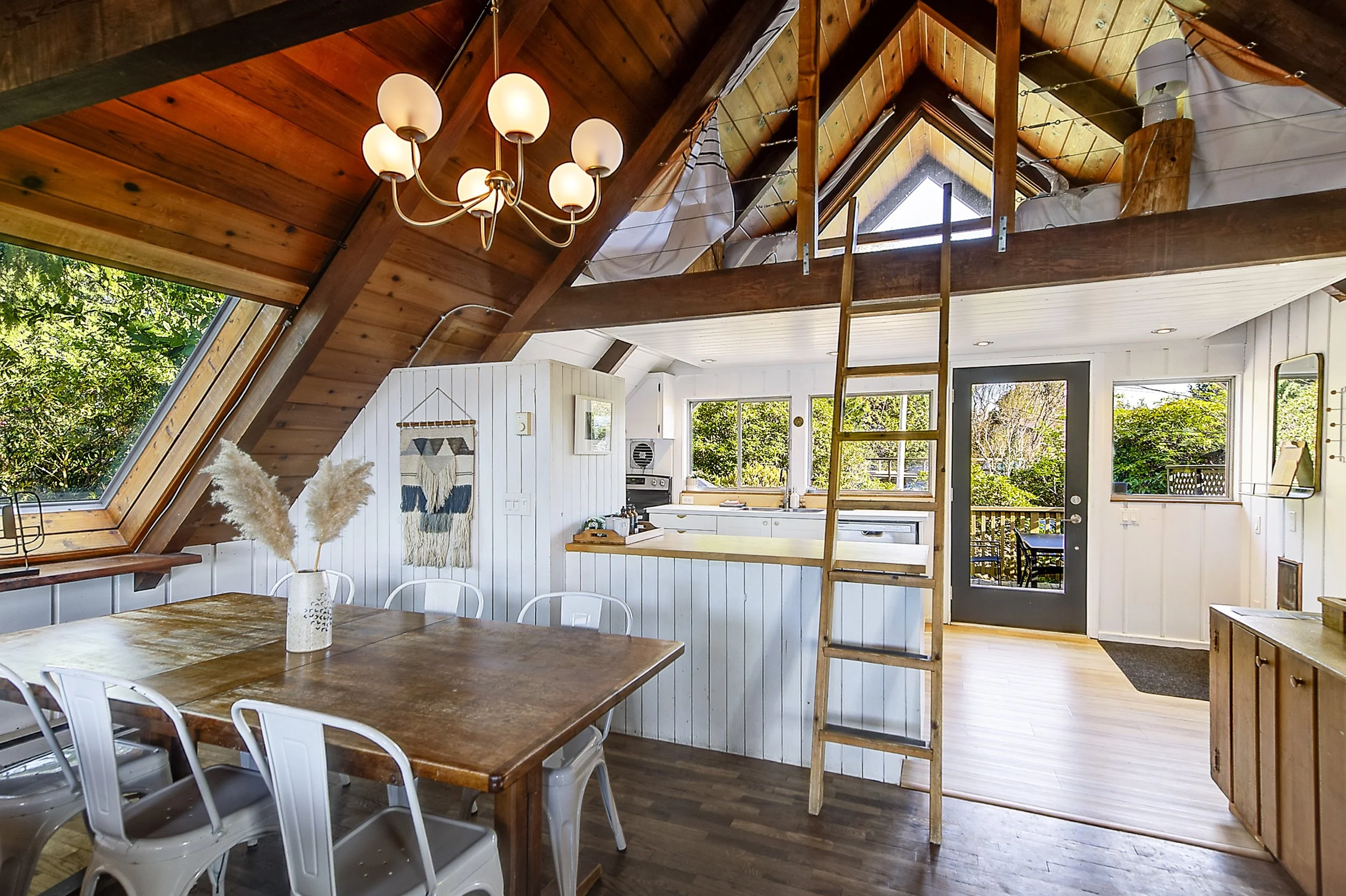 Interior view of a cozy, modern A-frame cabin with wood-paneled vaulted ceilings, a chandelier with multiple globes, white paneled walls, a wooden dining table with white chairs, and a loft accessible by a wooden ladder. Large windows and a glass doo