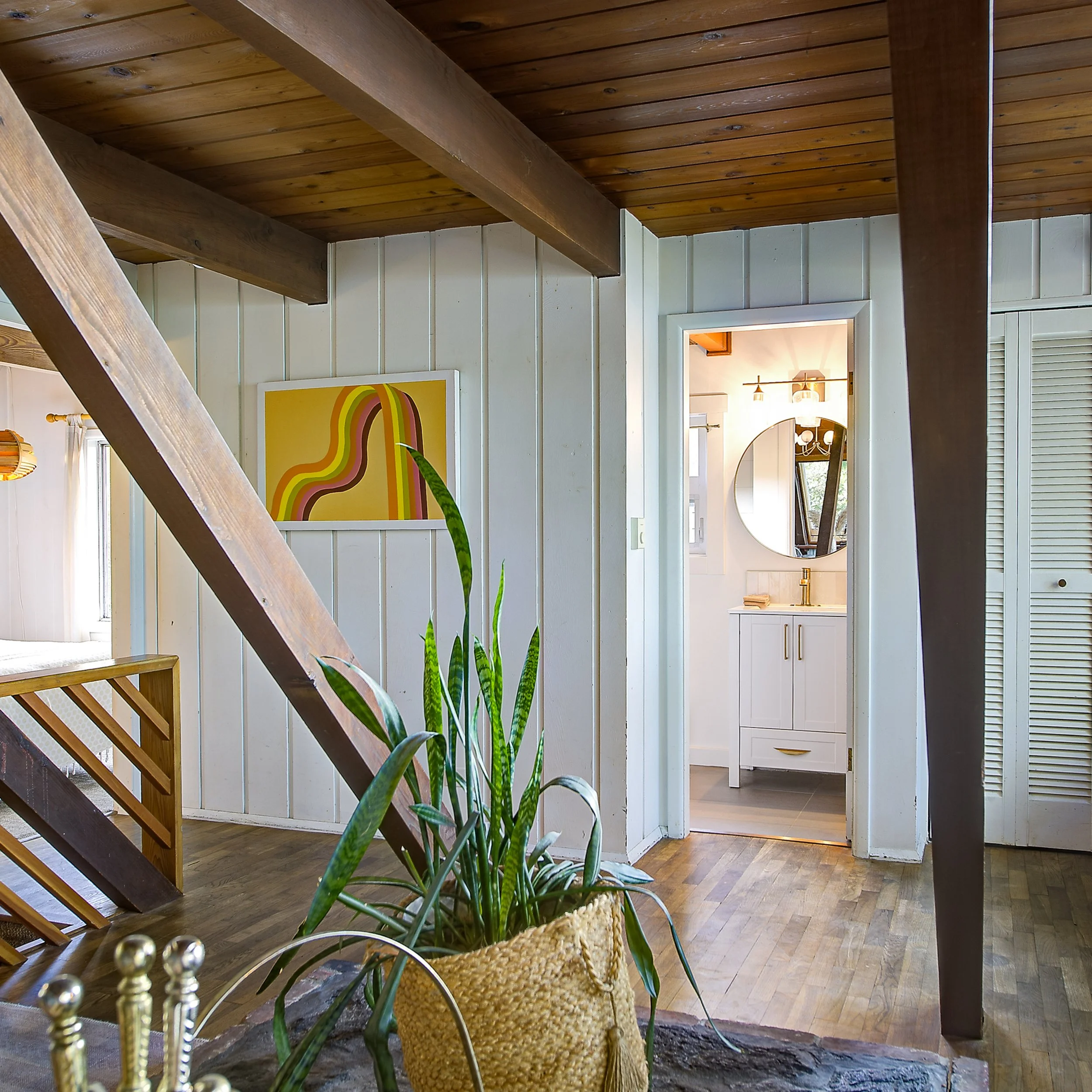 Interior of a house with wooden ceiling and white paneled walls, featuring a staircase, a potted plant, and a framed rainbow artwork.