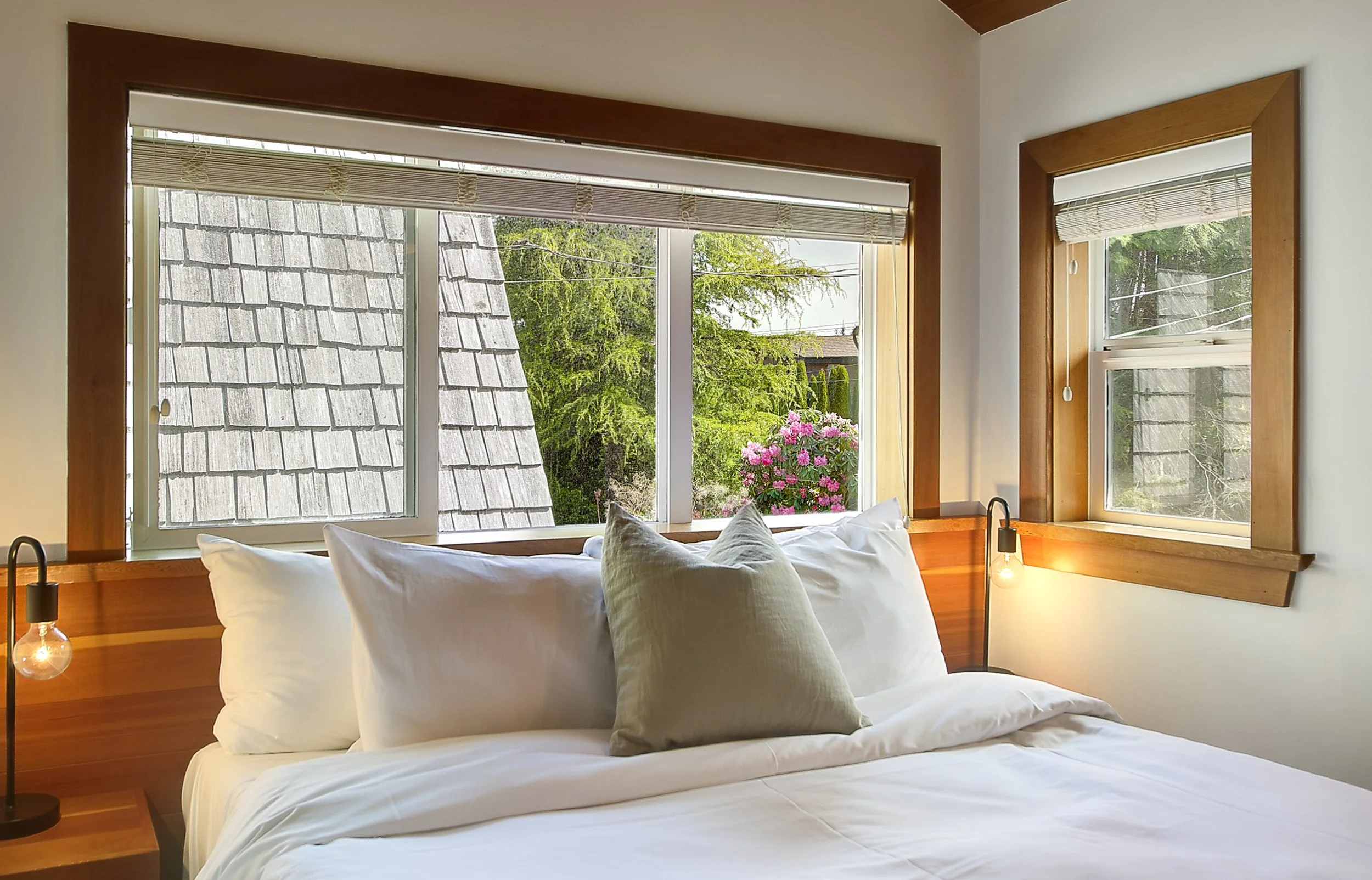 A bedroom with large windows showing a view of trees and a rooftop, with a bed in the foreground and pillows, a side table with a lamp, and a hanging light bulb.