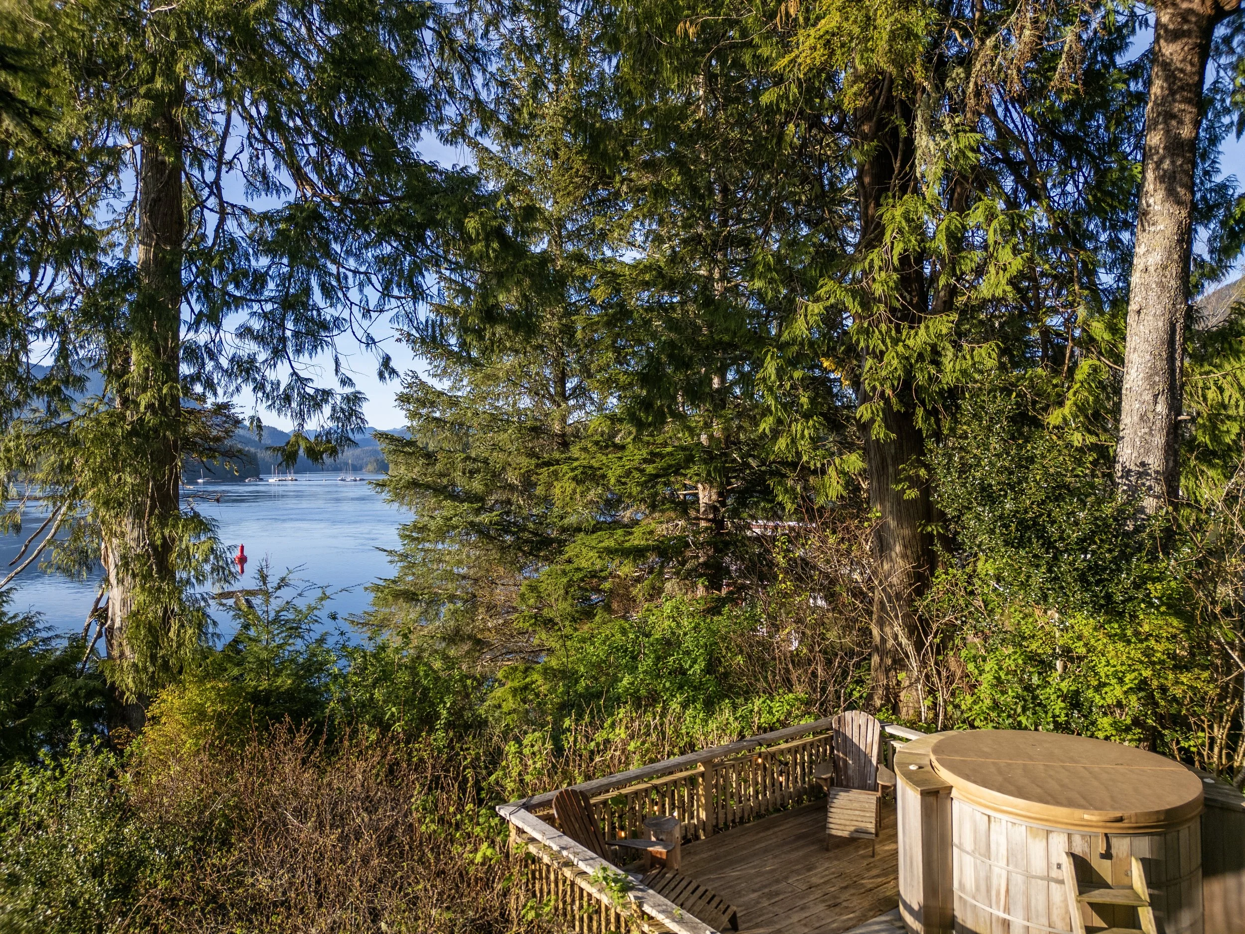 A lakeside view with lush green trees and a deck, featuring a hot tub and seating area with a table and chairs, overlooking a calm body of water with sailboats and a distant shoreline.