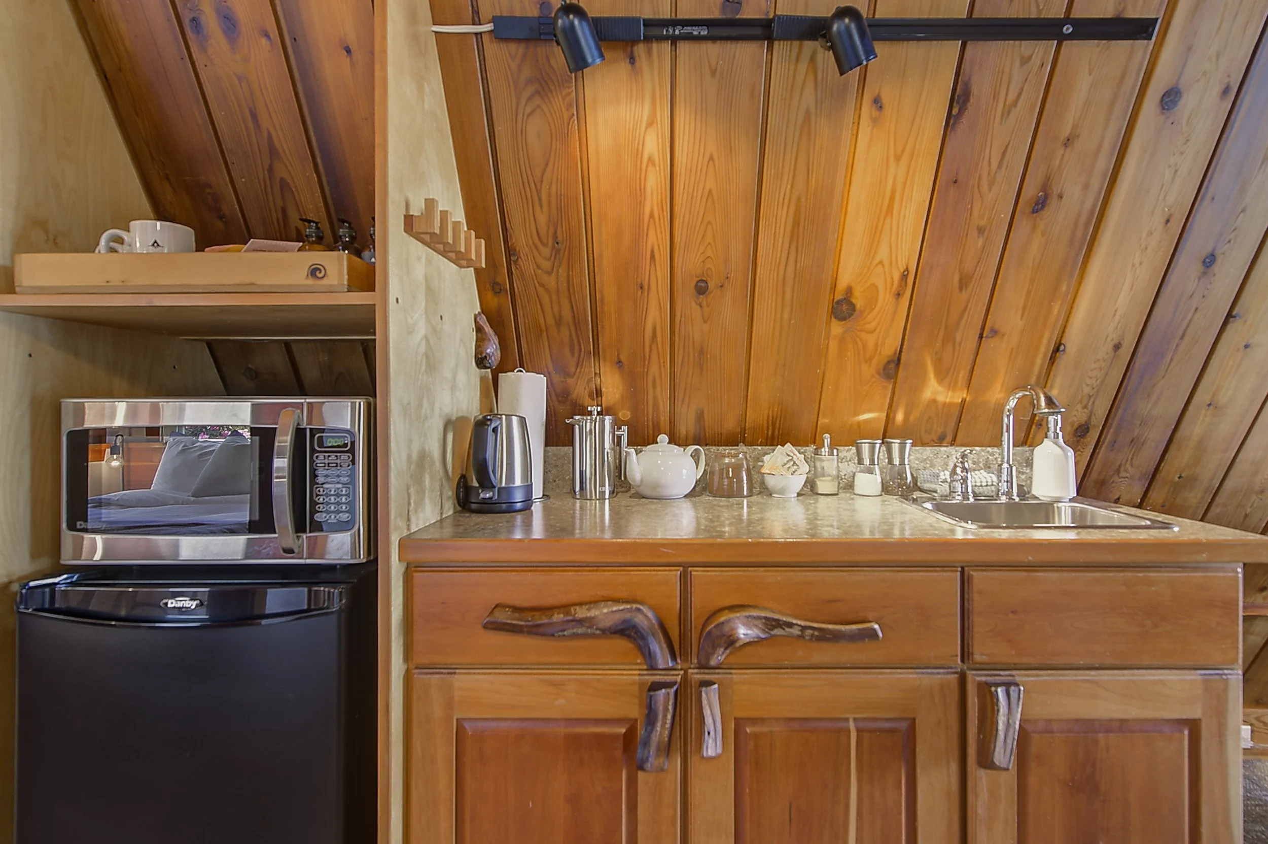 Wood-paneled kitchen with countertop appliances, tea kettle, mugs, and a sink. Wooden chairs are partially visible in front of the counter.