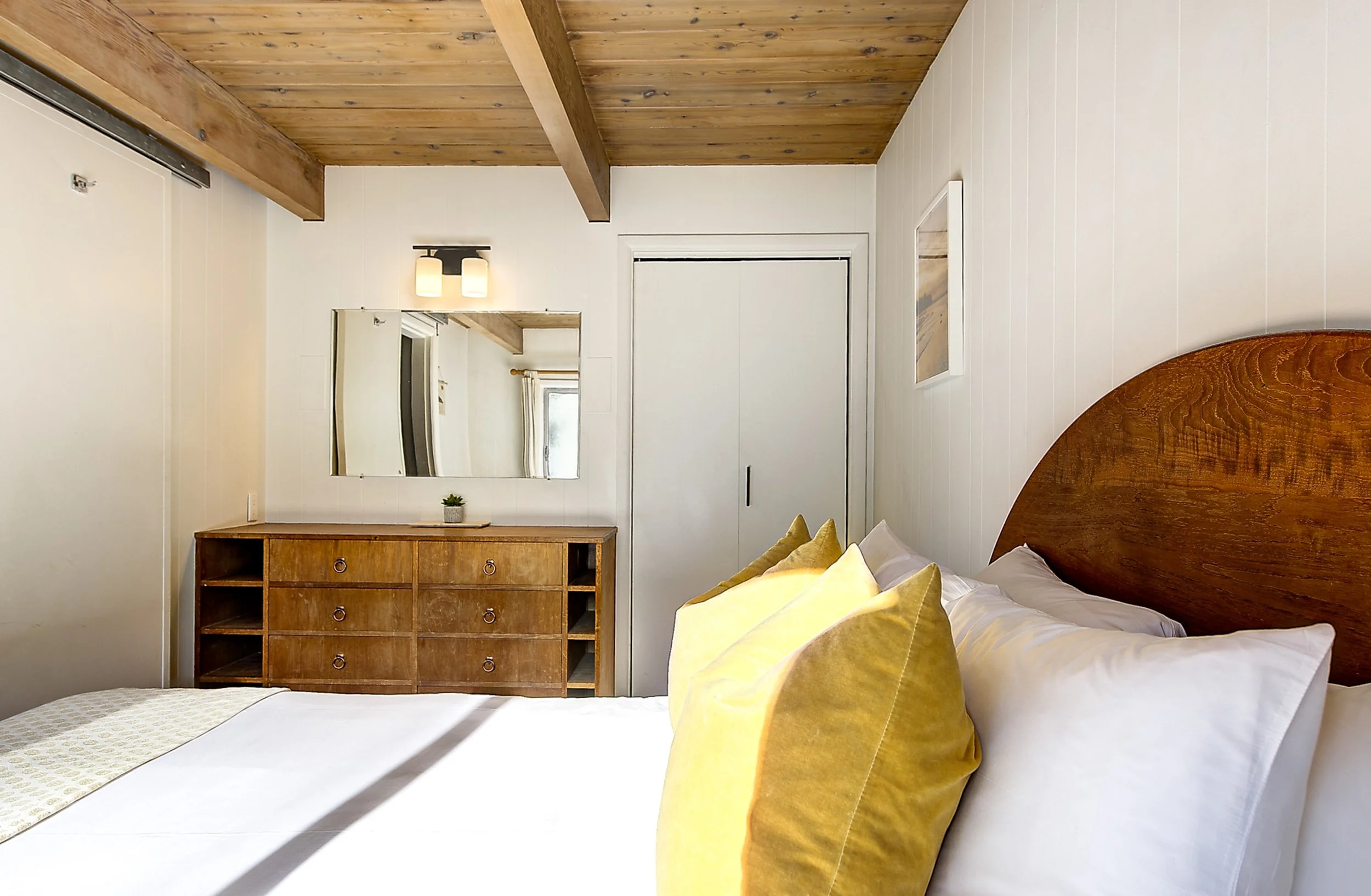 A cozy bedroom with a wooden bed frame and white bedding accented by yellow pillows. A wooden dresser with cubby holes is against the wall, and a mirror hangs above it. The ceiling is made of wood planks, and a light fixture with two lamps is mounted