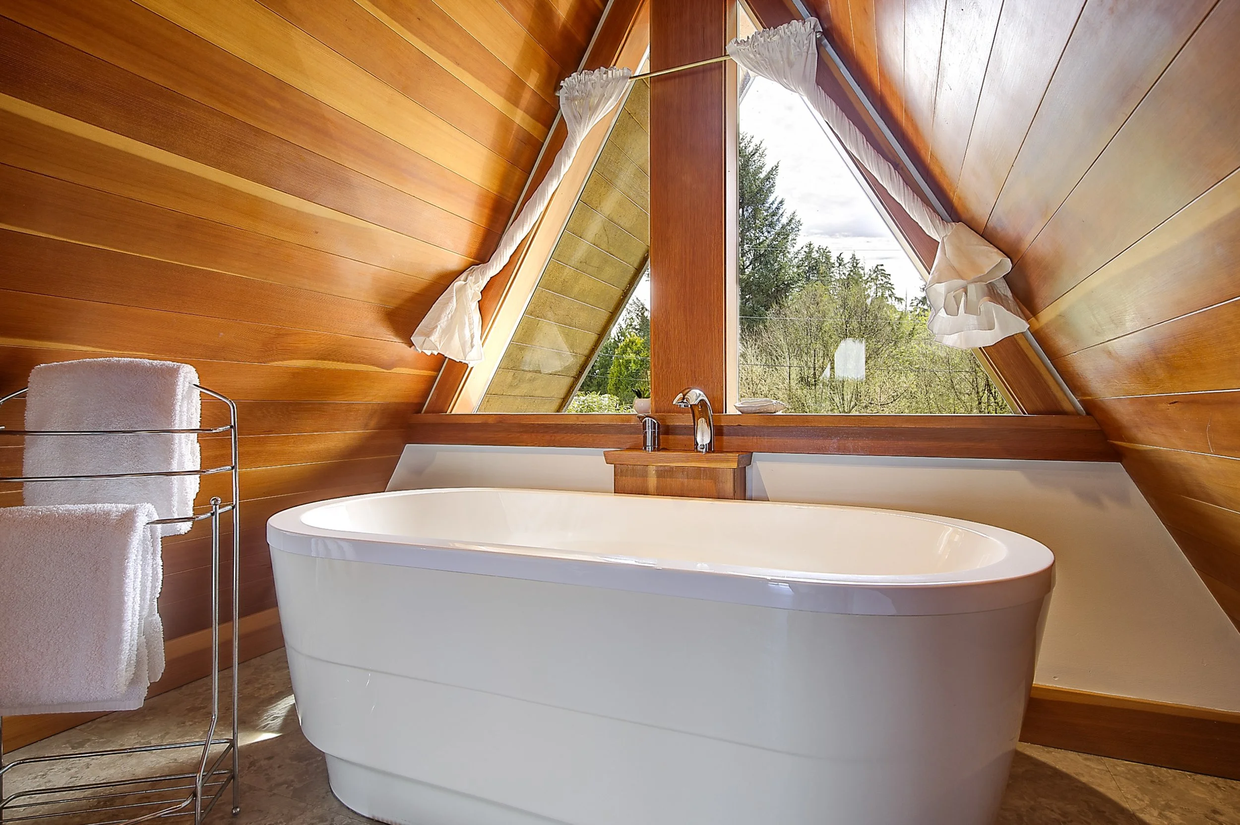 A bathroom with wood-paneled walls and ceiling, a large window with white curtains looking outside at trees, and a white bathtub.
