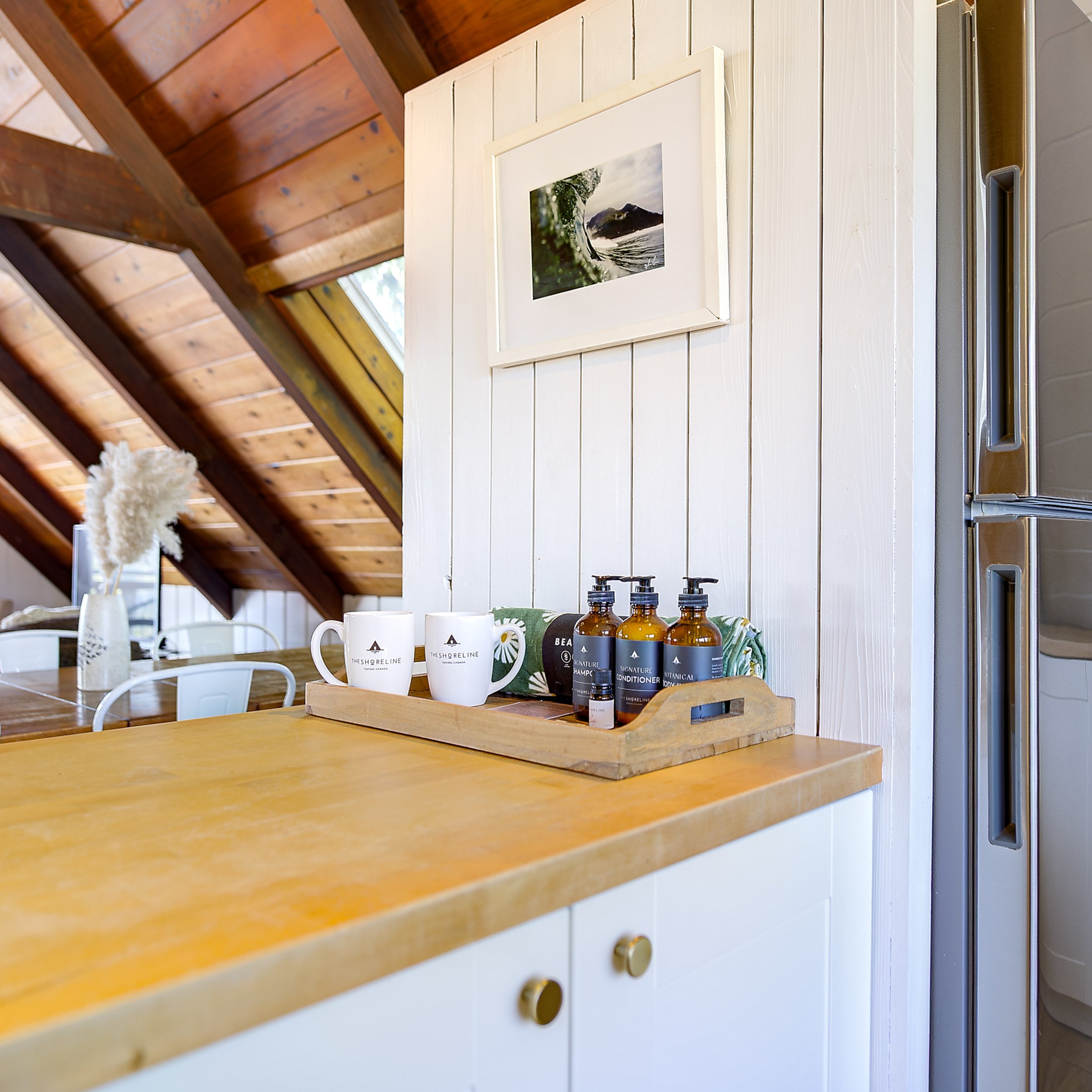 A wooden kitchen countertop with white wall paneling. Several bottles of toiletries are placed on the countertop, including shampoo and conditioner bottles. Two white coffee mugs are also on the countertop. A framed photograph of a wave crashing agai
