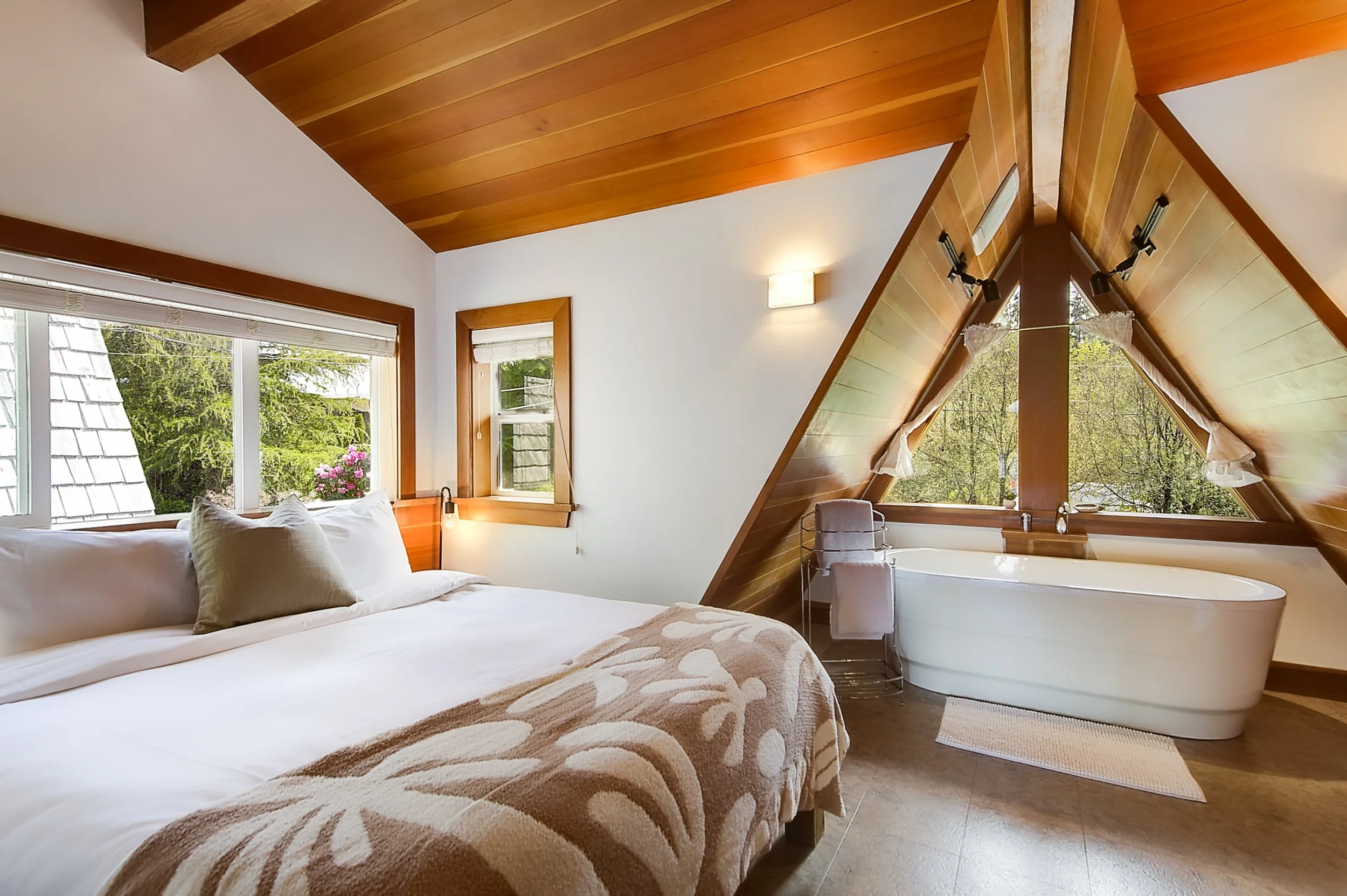 A cozy bedroom with wooden vaulted ceiling, large windows with white blinds, a comfortable bed with white sheets and beige patterned blanket, a beige pillow, and a corner with a bathtub and a chair, surrounded by greenery outside.