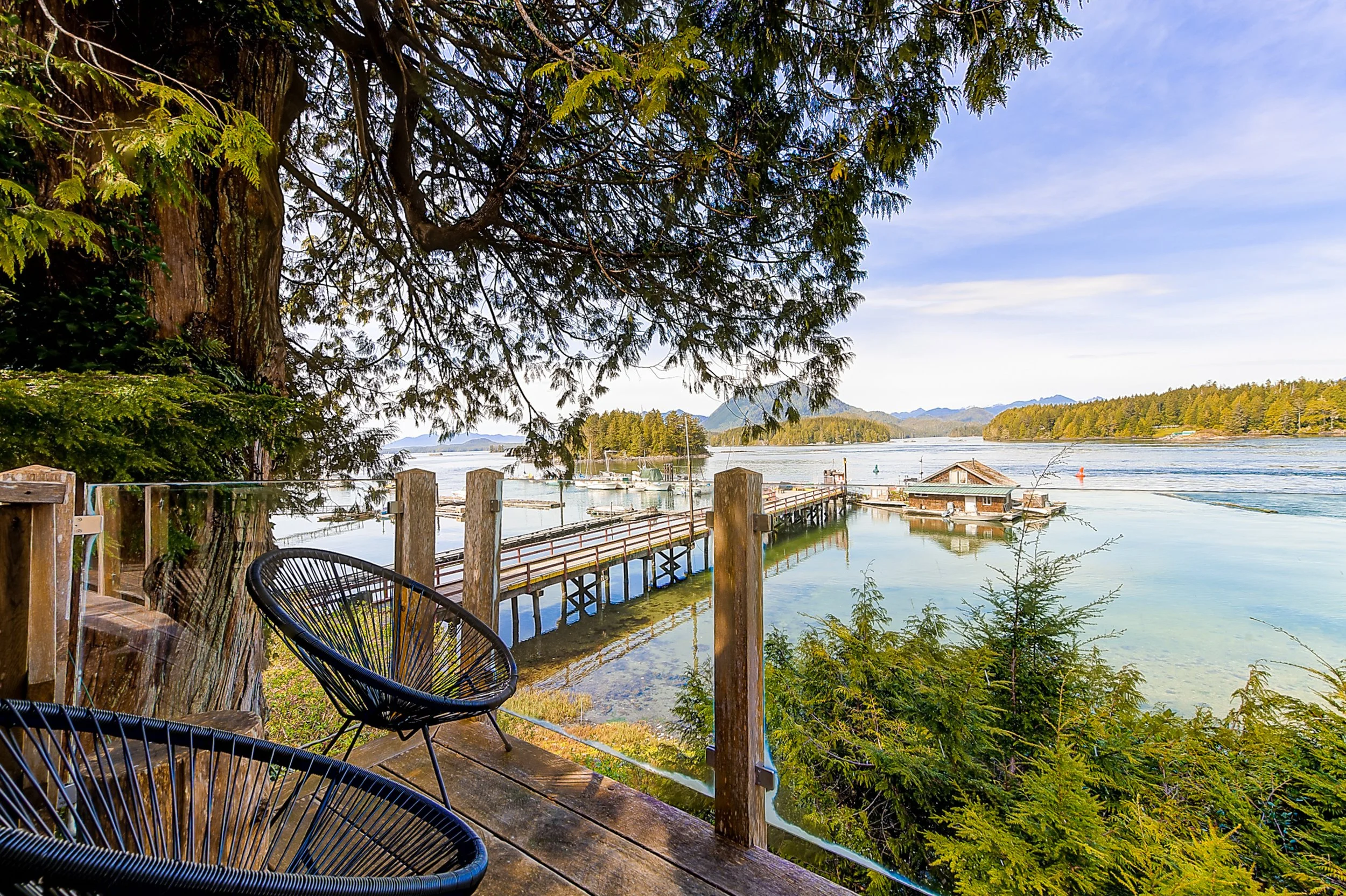 A peaceful lakeside scene viewed from a wooden deck with black chairs, tall green trees, calm water with boats, and a town with houses and distant mountains under a blue sky with some clouds.