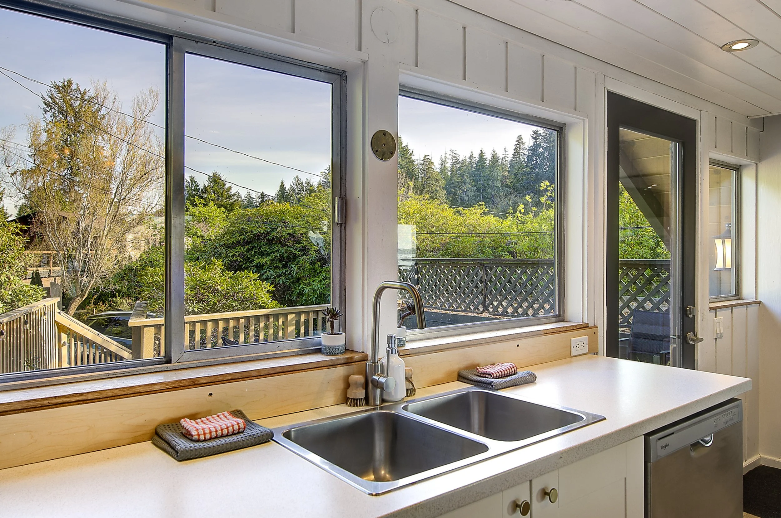 View from window showing trees, a wooden deck with railing, and a neighborhood in the distance outside a kitchen window.