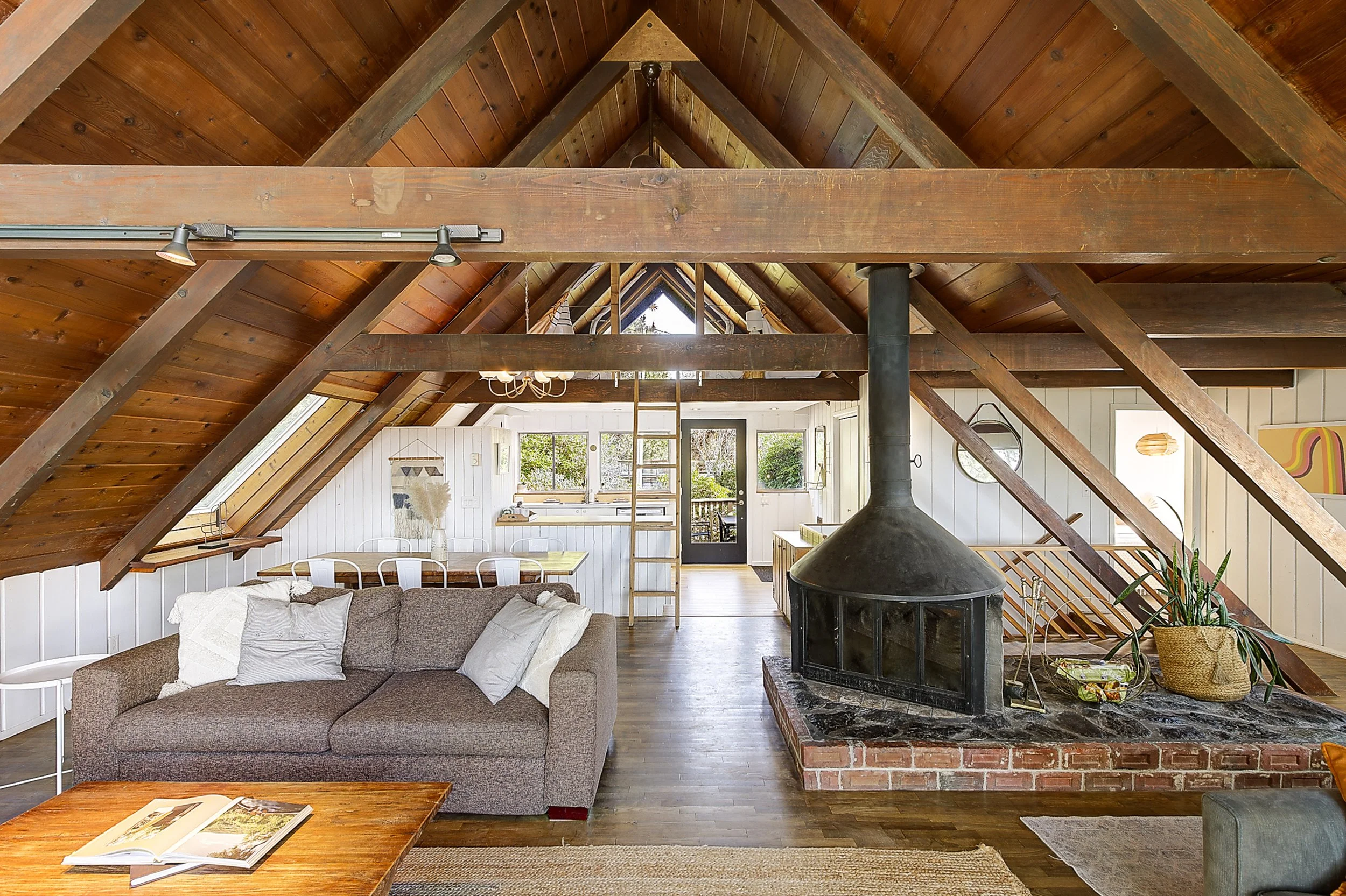 Living room with a brown sofa, white cushions, and a wooden coffee table with an open magazine. The room has a vaulted wood ceiling, a black stone fireplace, a dining area with a light-colored table and white chairs, and large windows letting in natu