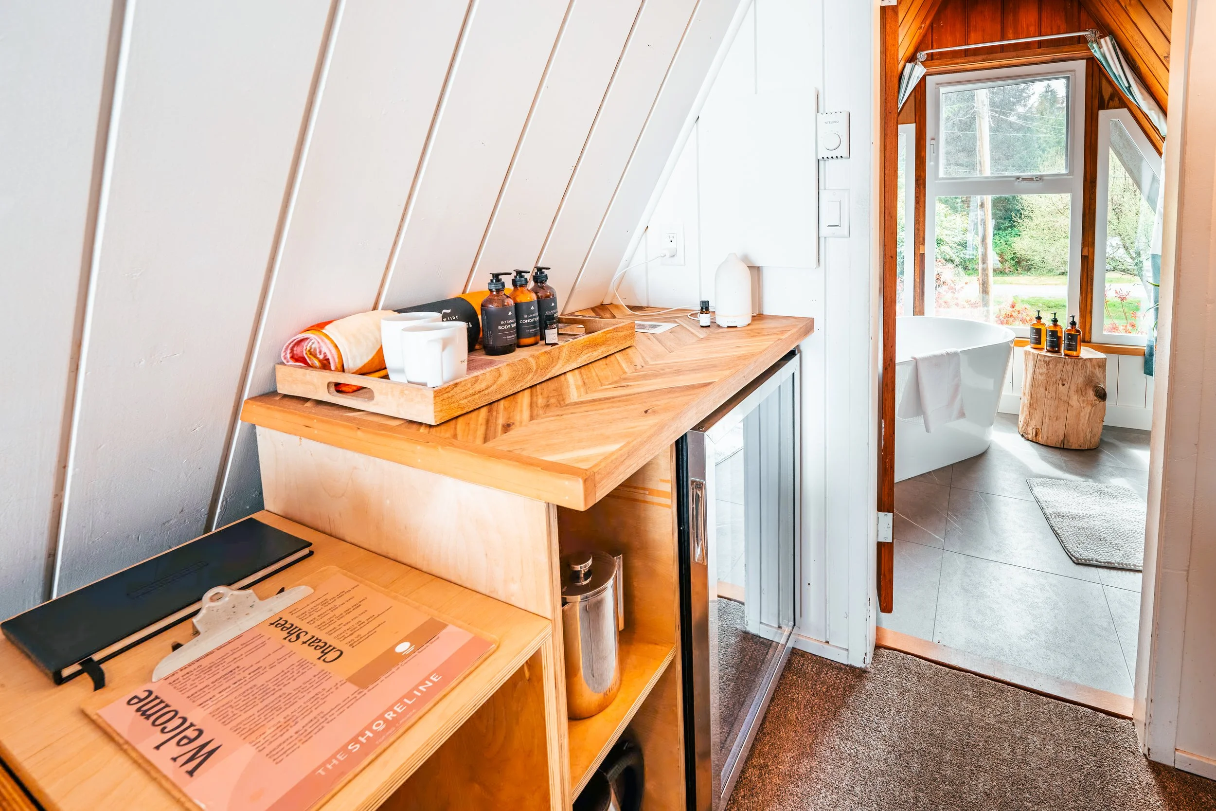A cozy bathroom with a wooden bathtub surrounded by large windows, and a sloped ceiling with white paneling. There is a small wooden side table with three bottles of soap, a towel, and a diffuser. The space includes a countertop with toiletries and a