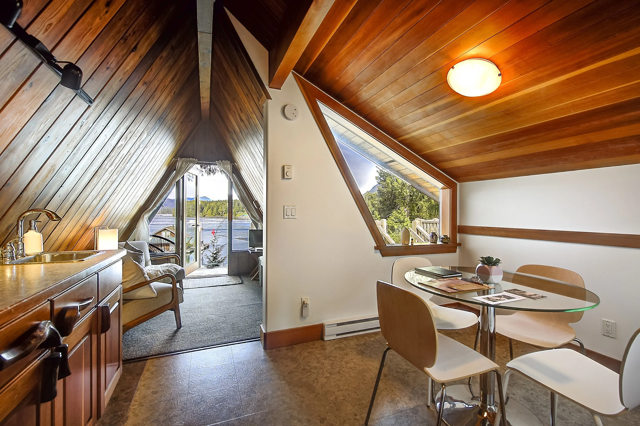 A cozy living space with a sloped wooden ceiling, a dining area with a glass table and beige chairs, and a view of trees and mountains through large windows.