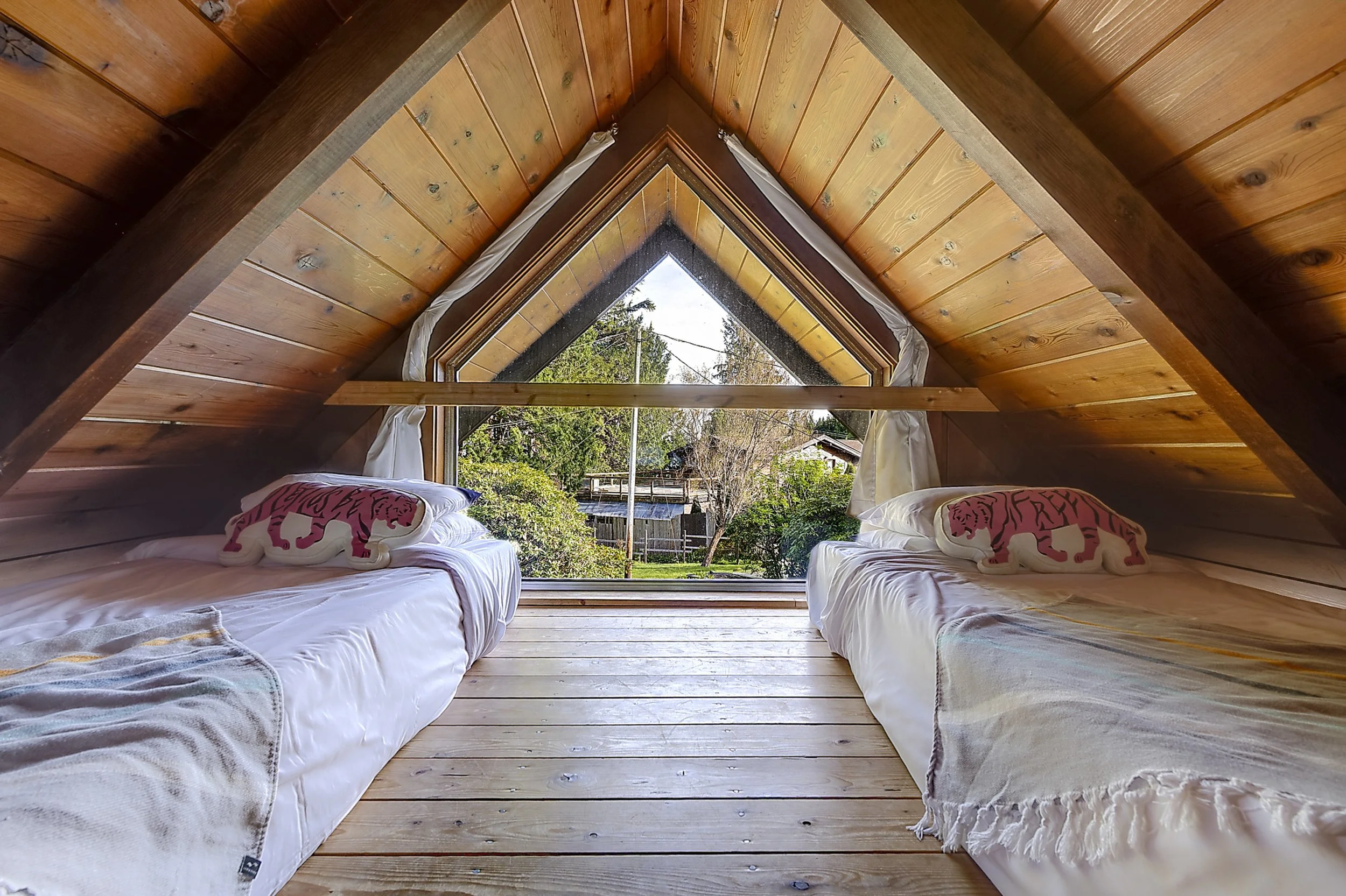 View of a cozy attic bedroom with twin beds adorned with slippers featuring pink tiger designs. The room has wooden walls and floors, a large triangular window with white curtains, and outdoor greenery visible through the window.