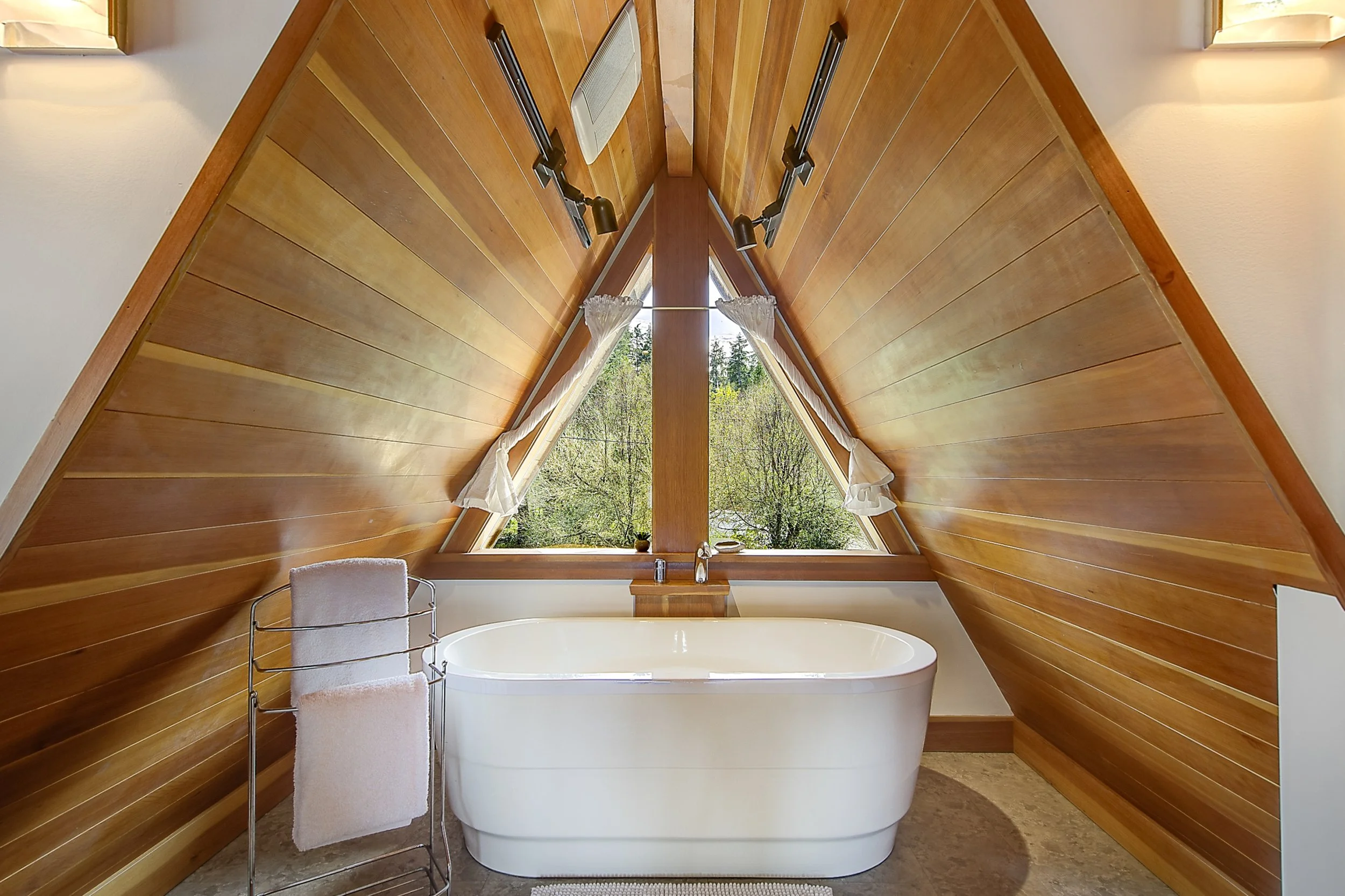 Bathroom with a freestanding bathtub below a large triangular window, surrounded by wood-paneled walls, with curtains on the window, a chair and towel near the tub.