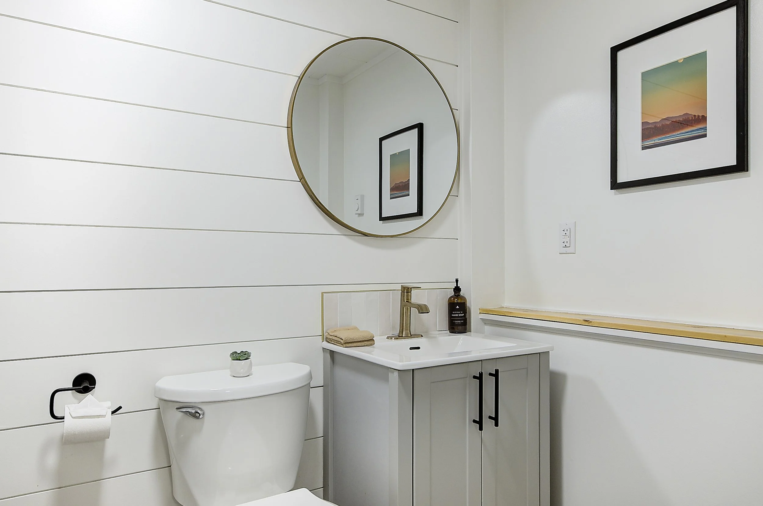 A modern bathroom featuring white shiplap walls, a round wall mirror with a thin gold frame, and a black framed artwork of a sunset over water. The vanity has a white sink with black handles, and a small potted succulent sits on a white surface. A bo