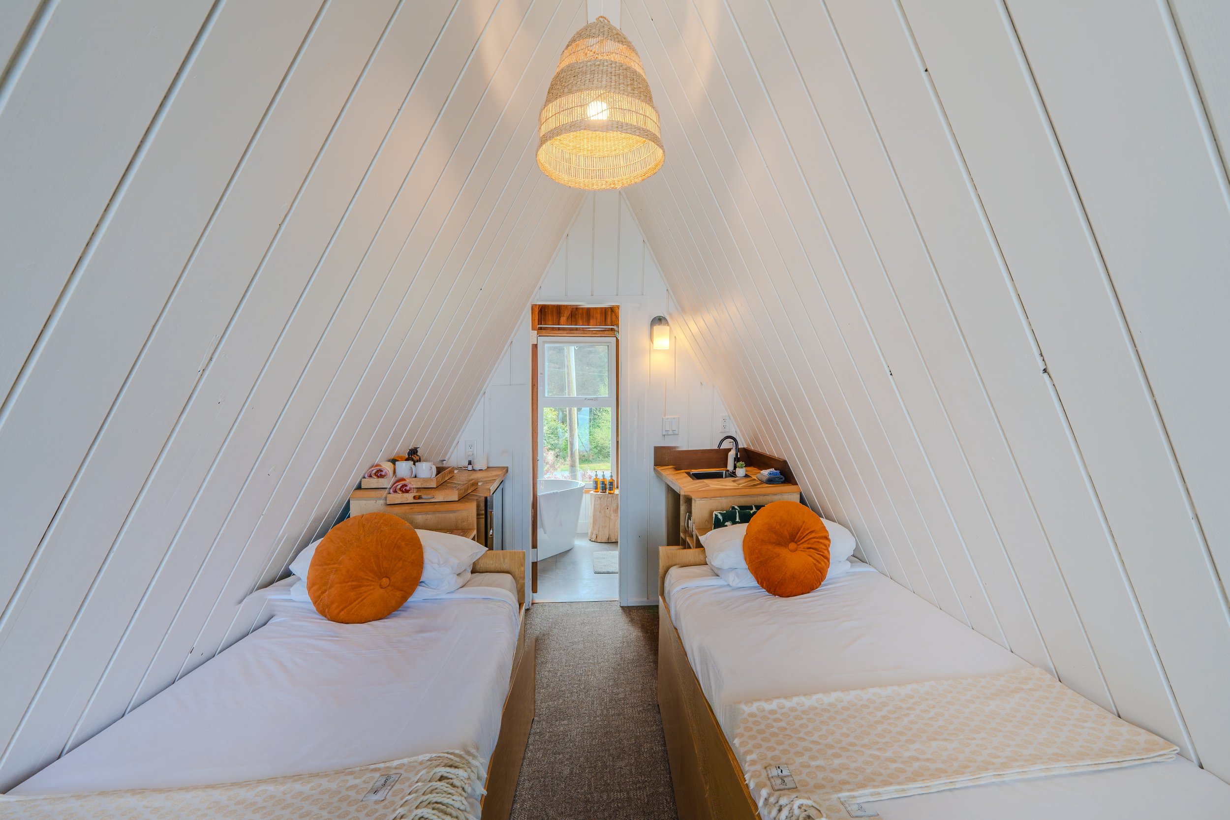 Two twin beds with white bedding and orange round pillows in an A-frame wooden cabin room. There are small wooden nightstands beside each bed with rolled towels on one. A window at the end of the room shows outdoor greenery. The ceiling and walls are