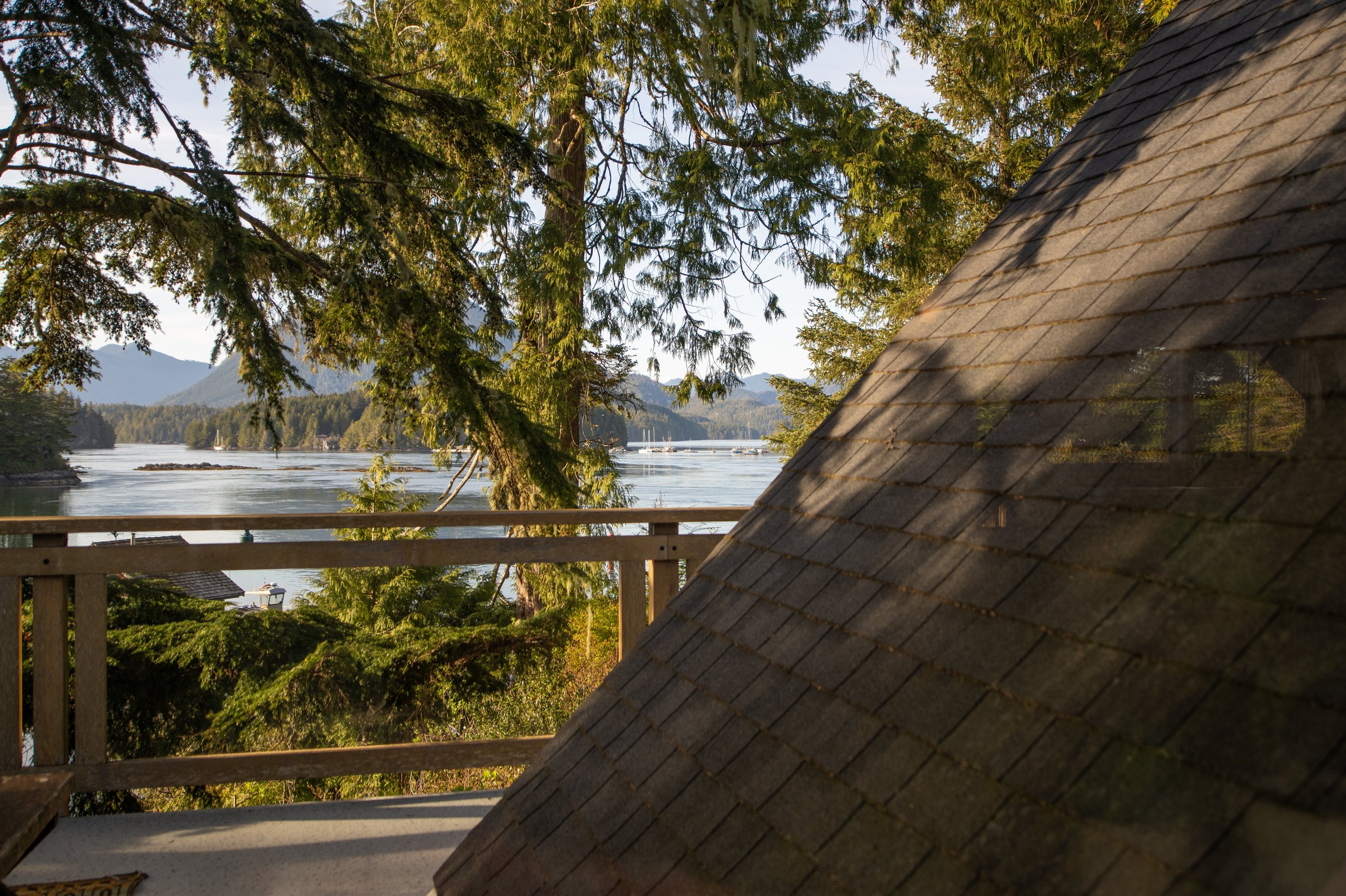 A view of a river surrounded by trees and mountains in the distance, partially obscured by the roof and railing of a wooden porch or balcony.