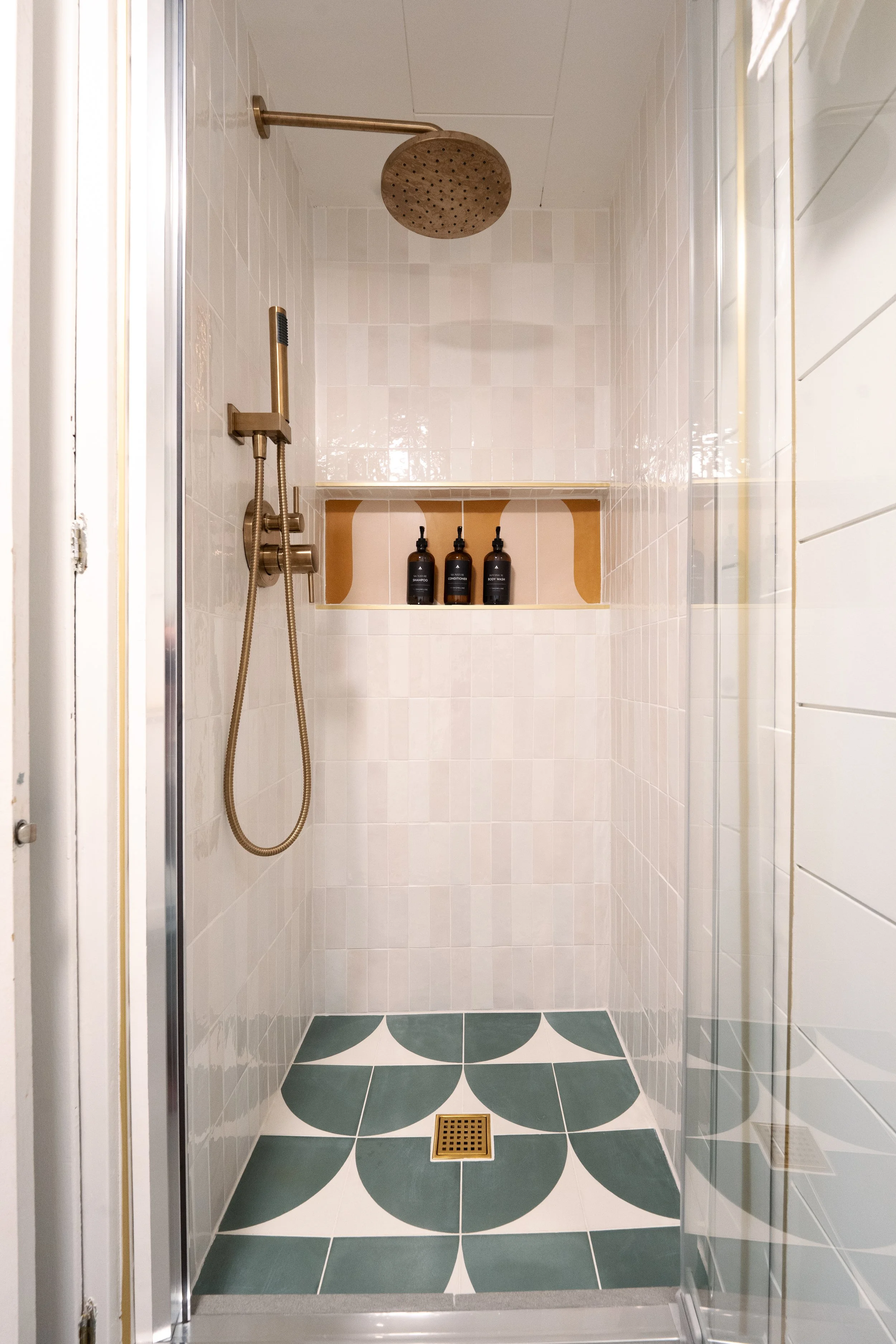 View of a small modern shower with white walls, a large bronze rainfall showerhead, a handheld shower attached to a wall-mounted bar, a niche with three black bottles, green and white patterned floor tiles, and a square drain.