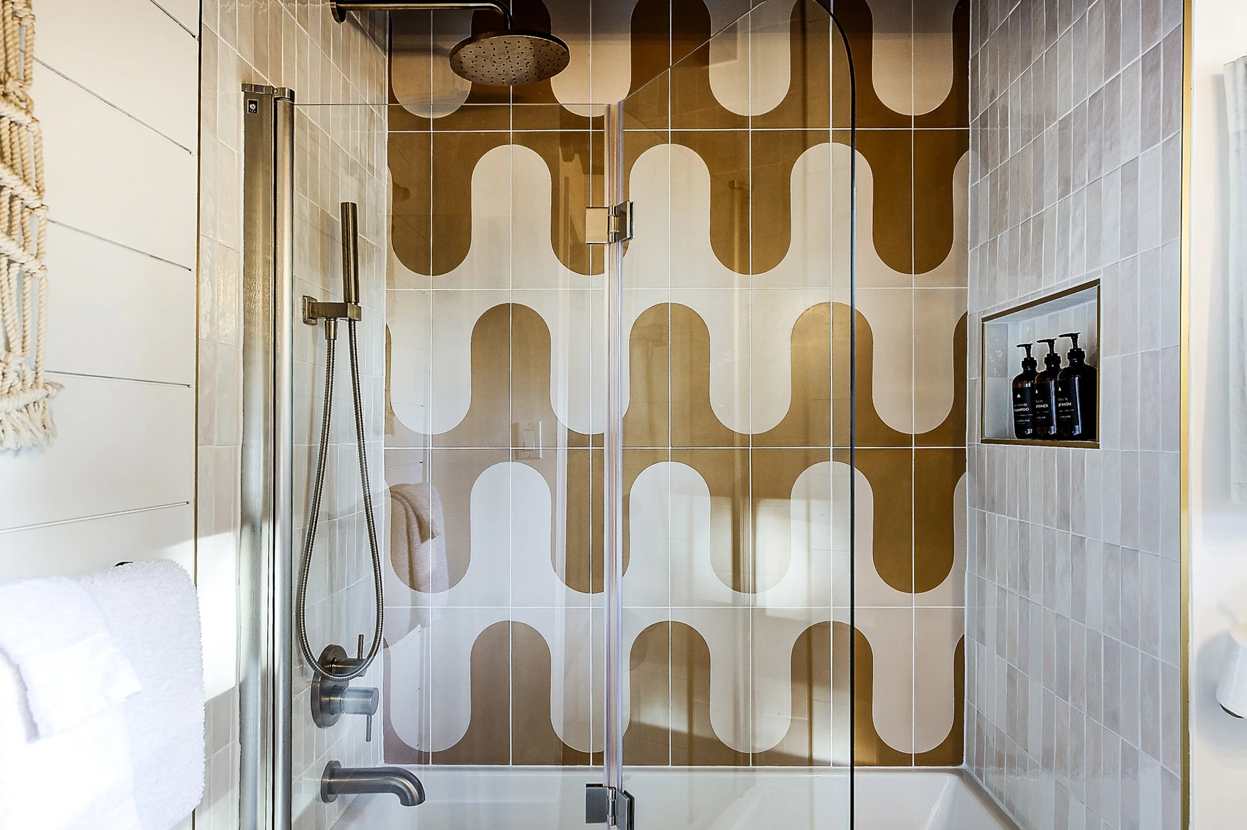 A modern shower with a glass door, brown and beige oval-patterned tiles on the wall, a round rainfall shower head, a handheld shower head, and black soap dispensers on a built-in shelf.
