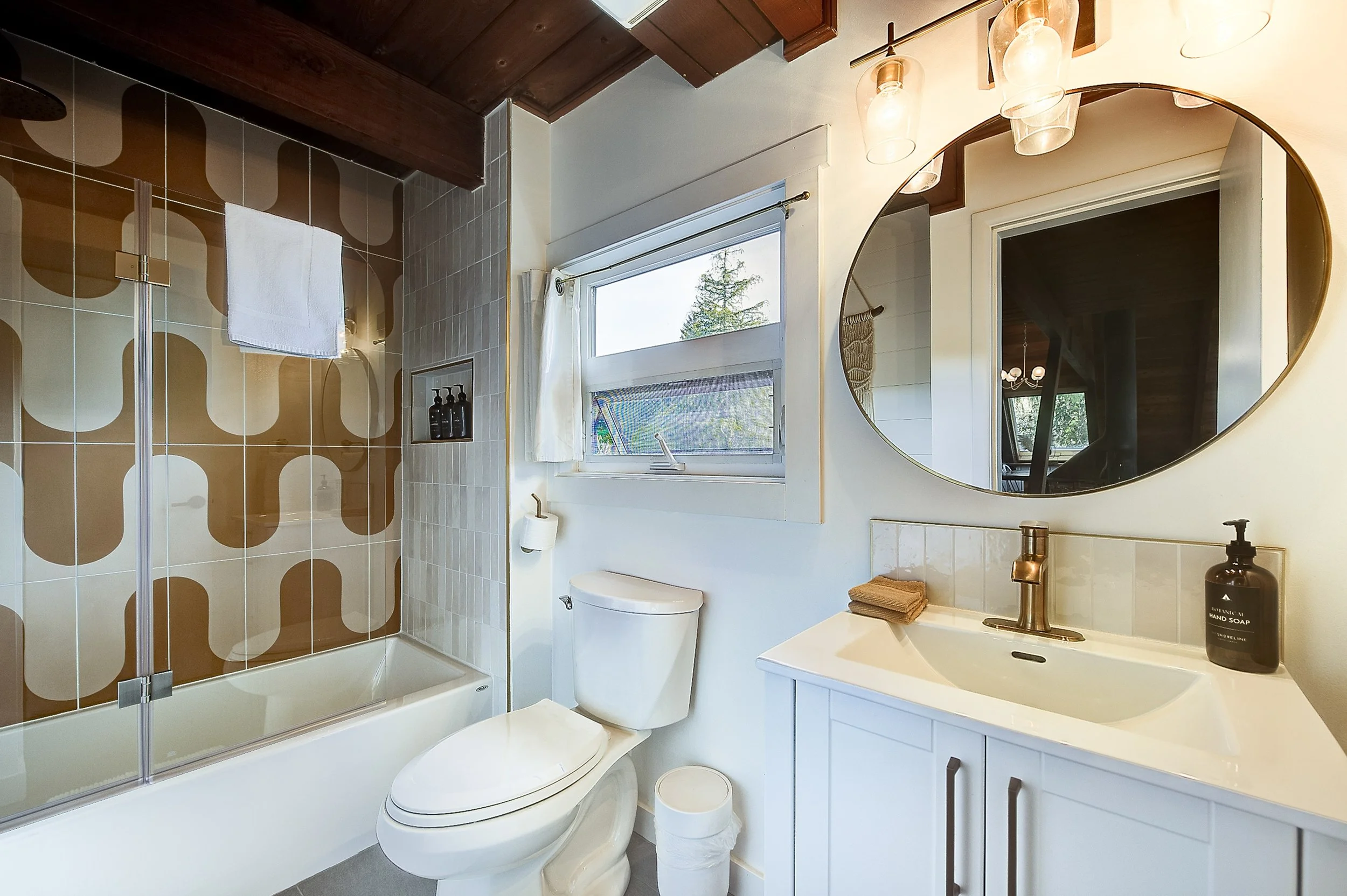 Bright bathroom with a walk-in shower, window with white curtains, white toilet, vanity with sink, round mirror, and modern light fixtures.