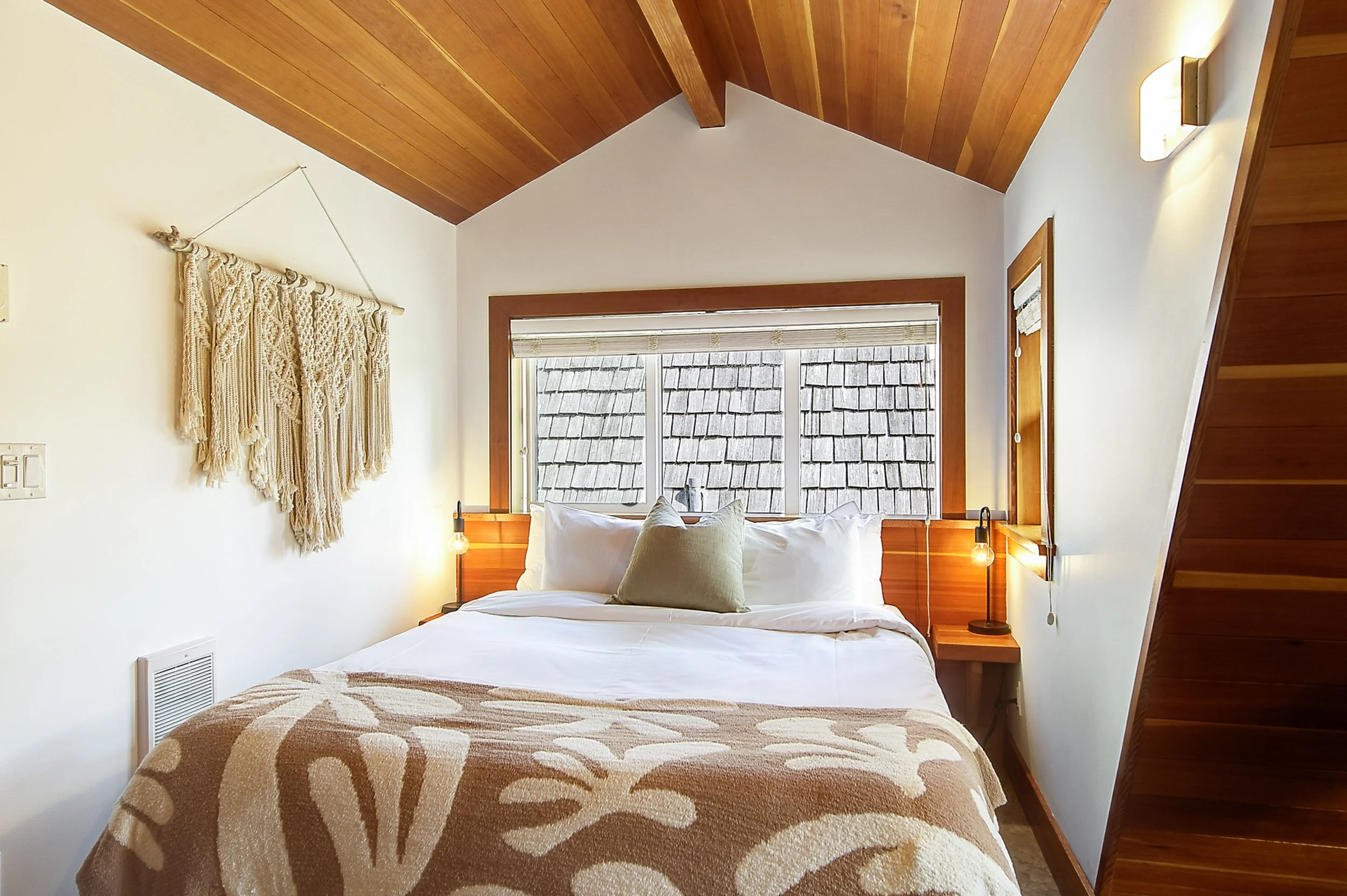 A cozy bedroom with a wooden ceiling, a large window with blinds, a bed with white linens and decorative pillows, a macramé wall hanging, and a wooden nightstand with a lamp.