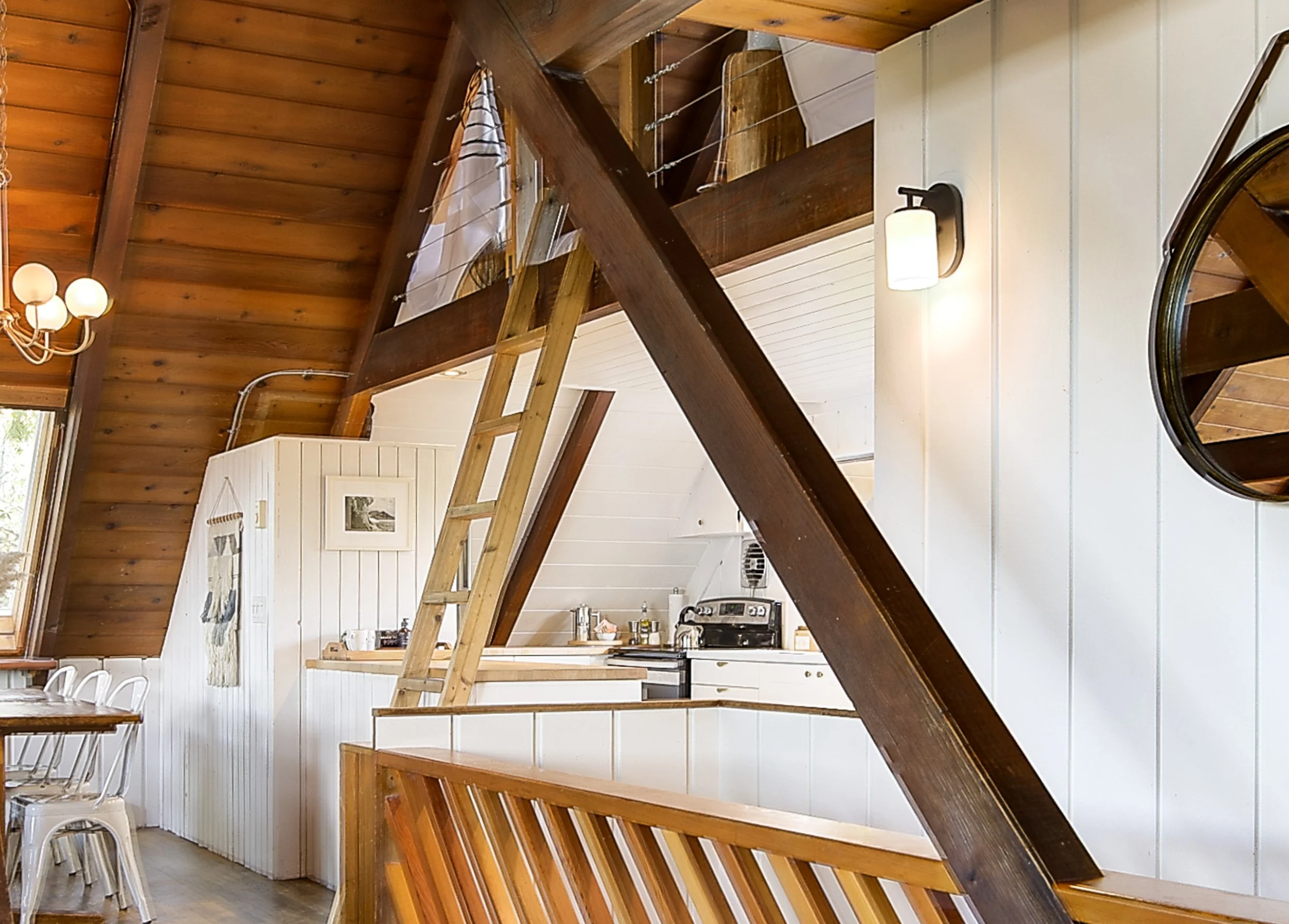 Interior view of a cozy wooden cabin kitchen with a loft area accessible by a wooden ladder, white paneled walls, and warm lighting.