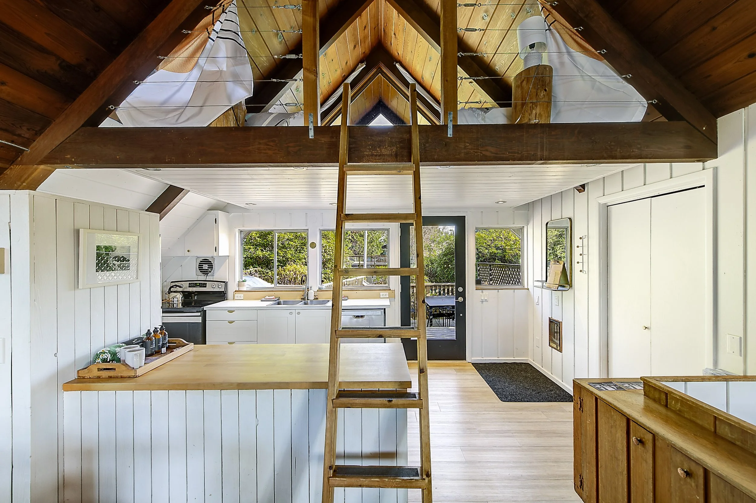 View of a cozy kitchen with white walls and wooden accents in a house with a loft. A wooden ladder leads to the loft area with exposed wooden beams and a sloped ceiling. Large windows provide natural light and a view of trees outside.