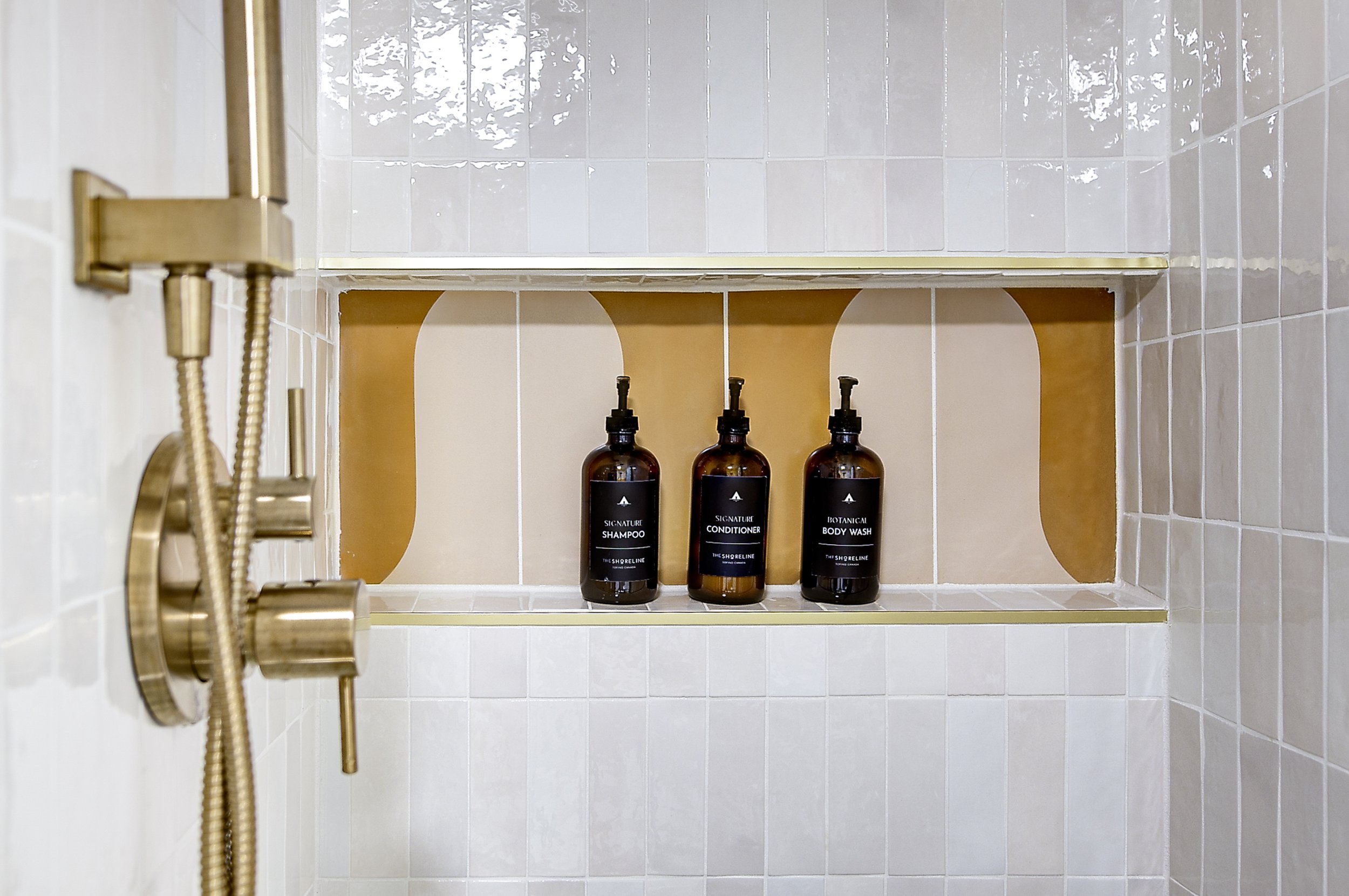 Bathroom shower shelf holding three bottles labeled shampoo, conditioner, and body wash in a tiled shower with a gold-colored shower fixture and white tiles.