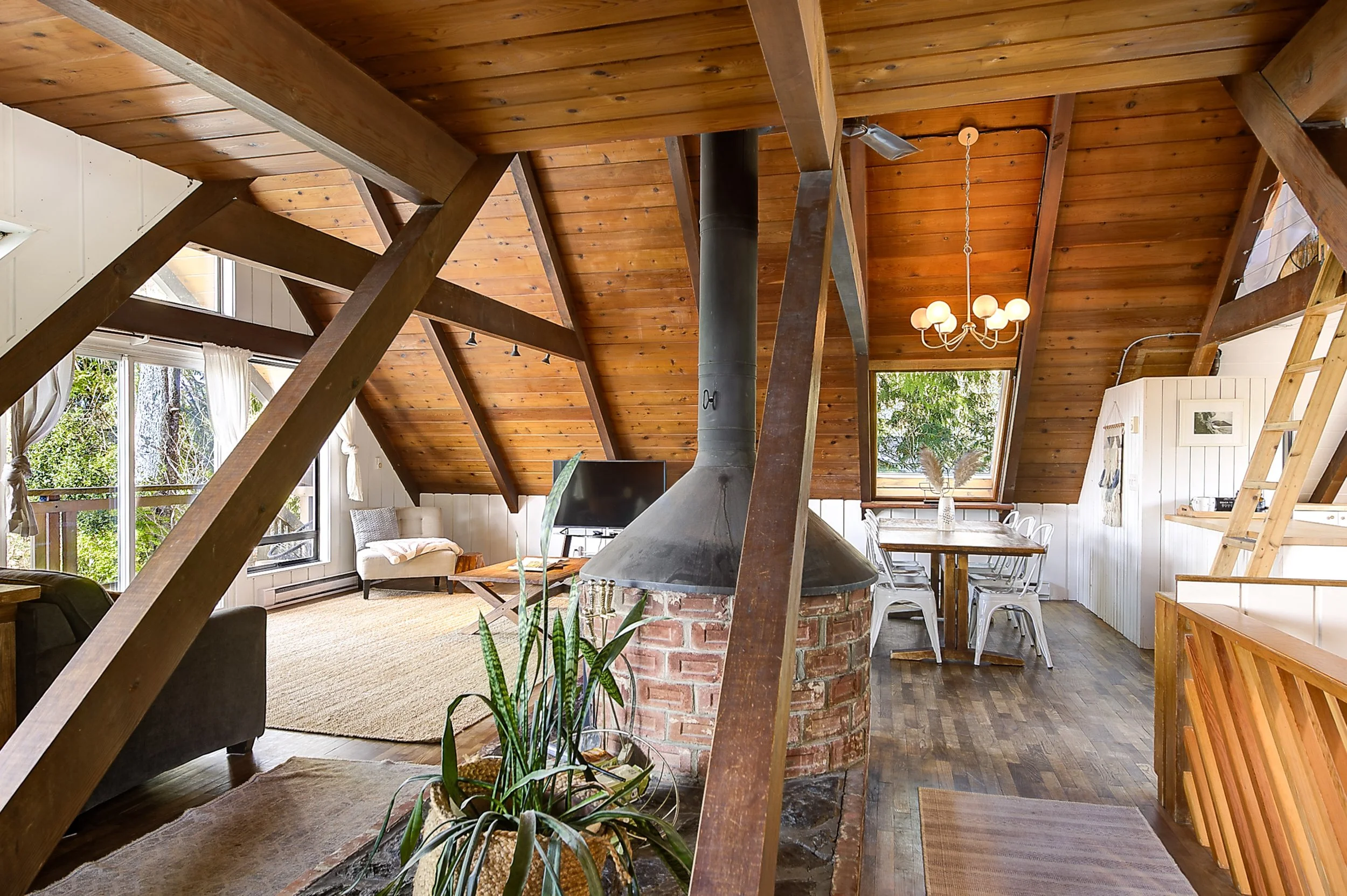 Interior of a cozy living space with a wood-paneled ceiling, a chandelier, a sofa with cushions, large windows with curtains, and a dining area with a table and chairs. Potted plants are in the foreground, and a black chimney pipe extends through the