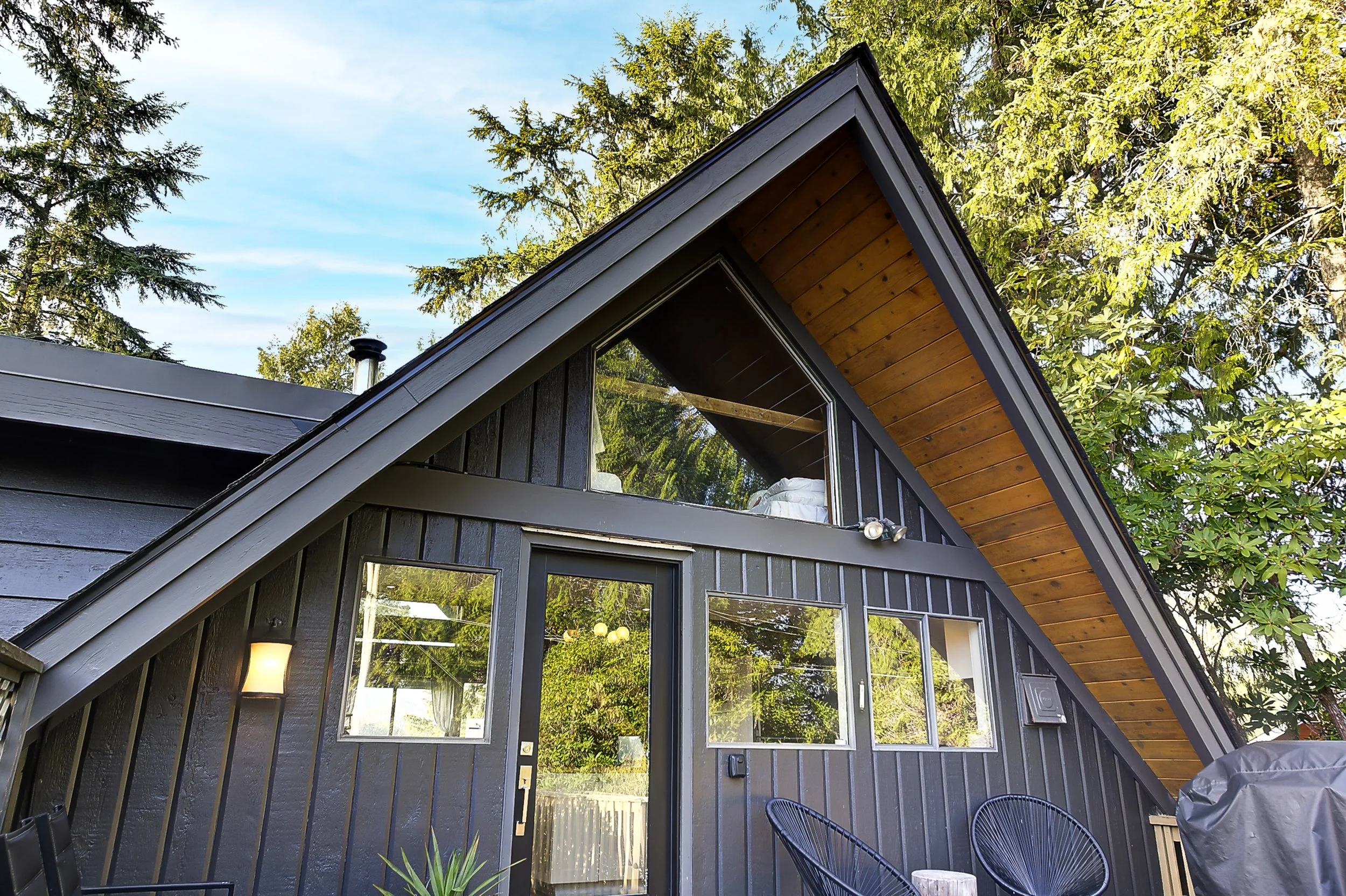 Close-up of modern house with dark gray exterior, large windows, front door, and outdoor seating area with black chairs and a table under the eaves; trees and sky in the background.