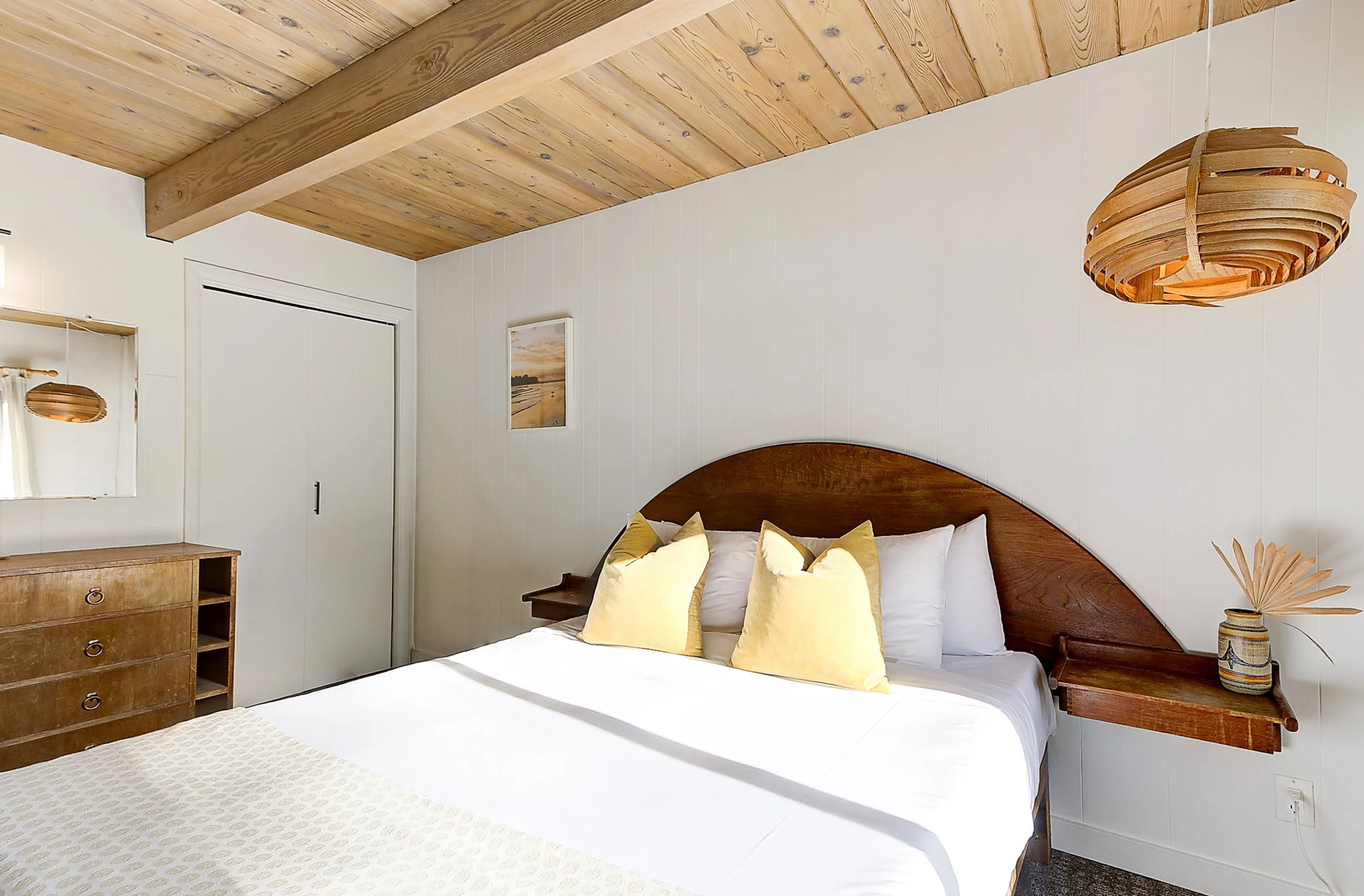 A bedroom with a wooden ceiling and white walls. There are several yellow pillows on a bed with white bedding, a wooden bedside table, a decorative vase with dried plants, and a framed sunset photo on the wall.
