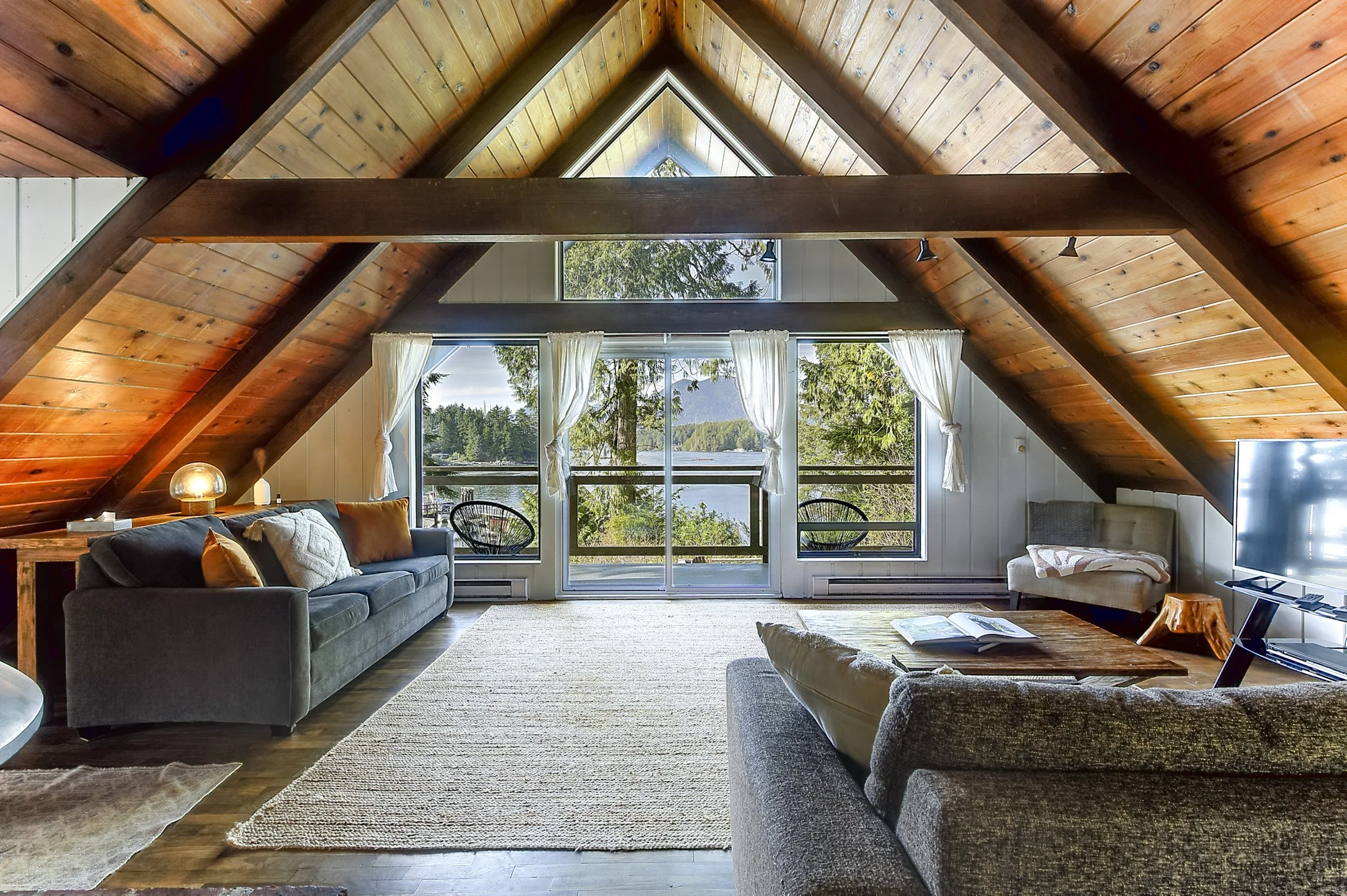 A spacious living room with wooden vaulted ceilings and large windows overlooking trees and water. The room is furnished with a gray sofa, a beige armchair, a black woven chair, and a wooden coffee table. Curtains frame the windows, and there is a ce