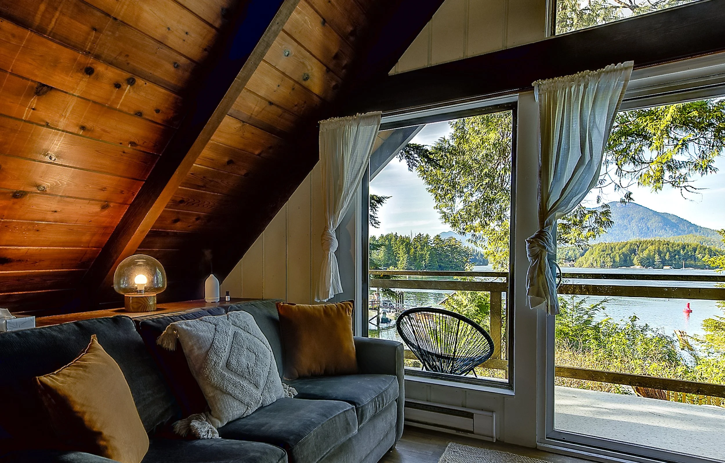 A cozy living room with a black leather sofa adorned with various pillows, wooden ceiling beams, a window with white curtains, and a scenic view of trees and a lake outside.