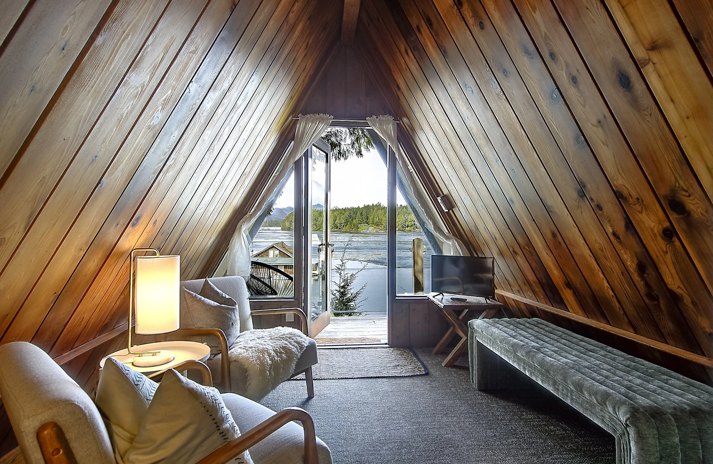 Room with wooden-paneled walls and A-frame ceiling, featuring a large window with a view of a lake, trees, and distant mountains. Inside, there is a beige couch with pillows, a white furry ottoman, a small side table with a lamp, a television, and a 