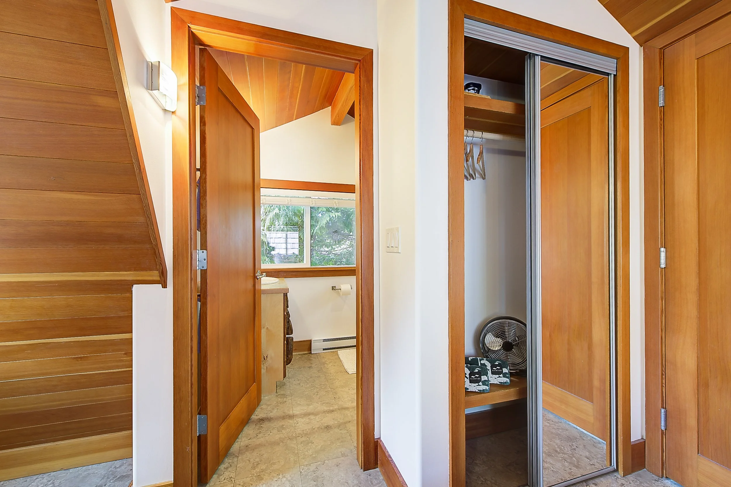View inside a house showing a wooden door leading to a bathroom with a window, and a closet with sliding mirror doors, a fan, and clothes hanging inside.