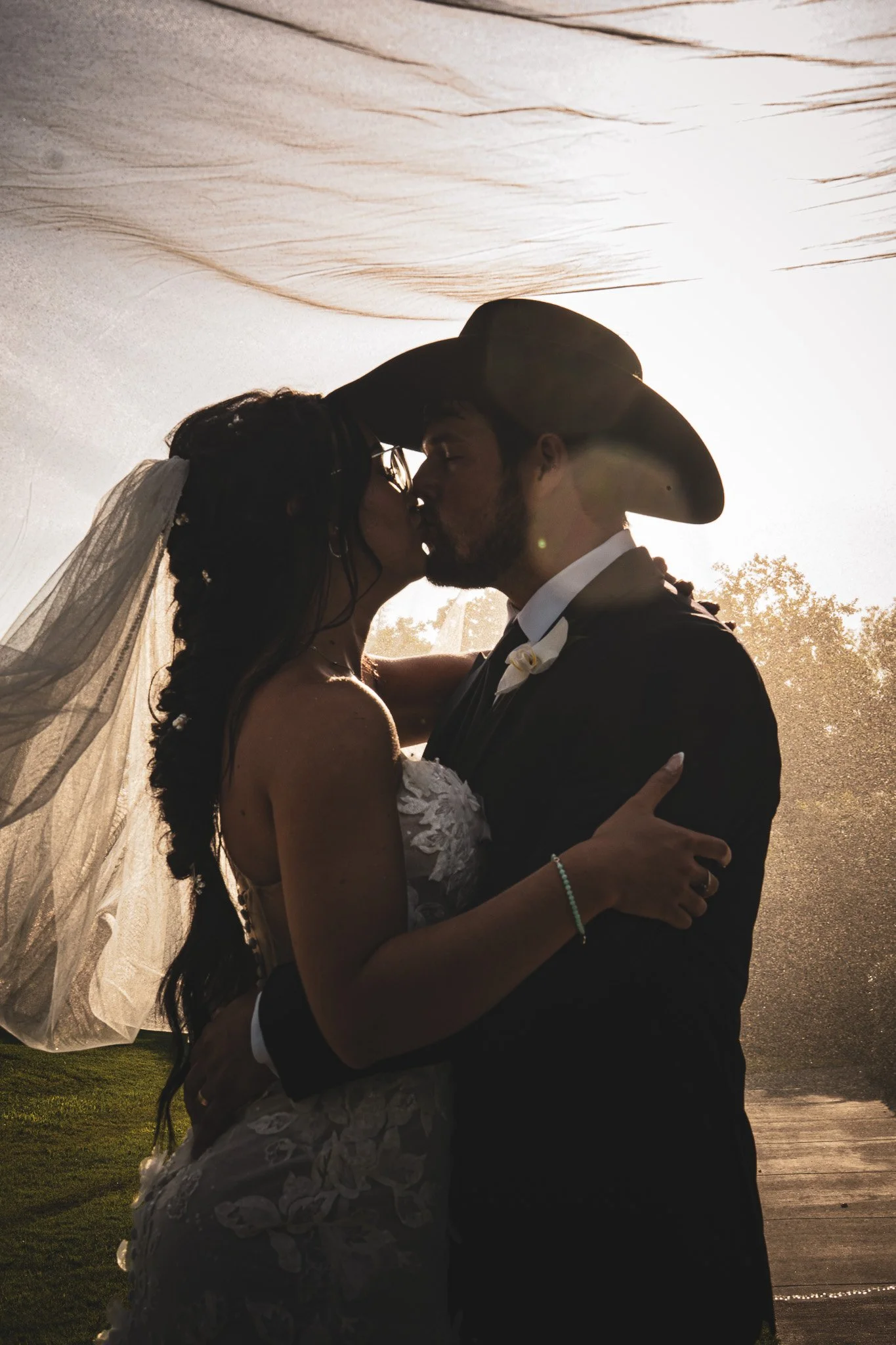 A couple kissing on their wedding day, with the bride wearing a veil and lace dress, and the groom wearing a cowboy hat, outdoors in front of the sun setting behind trees.