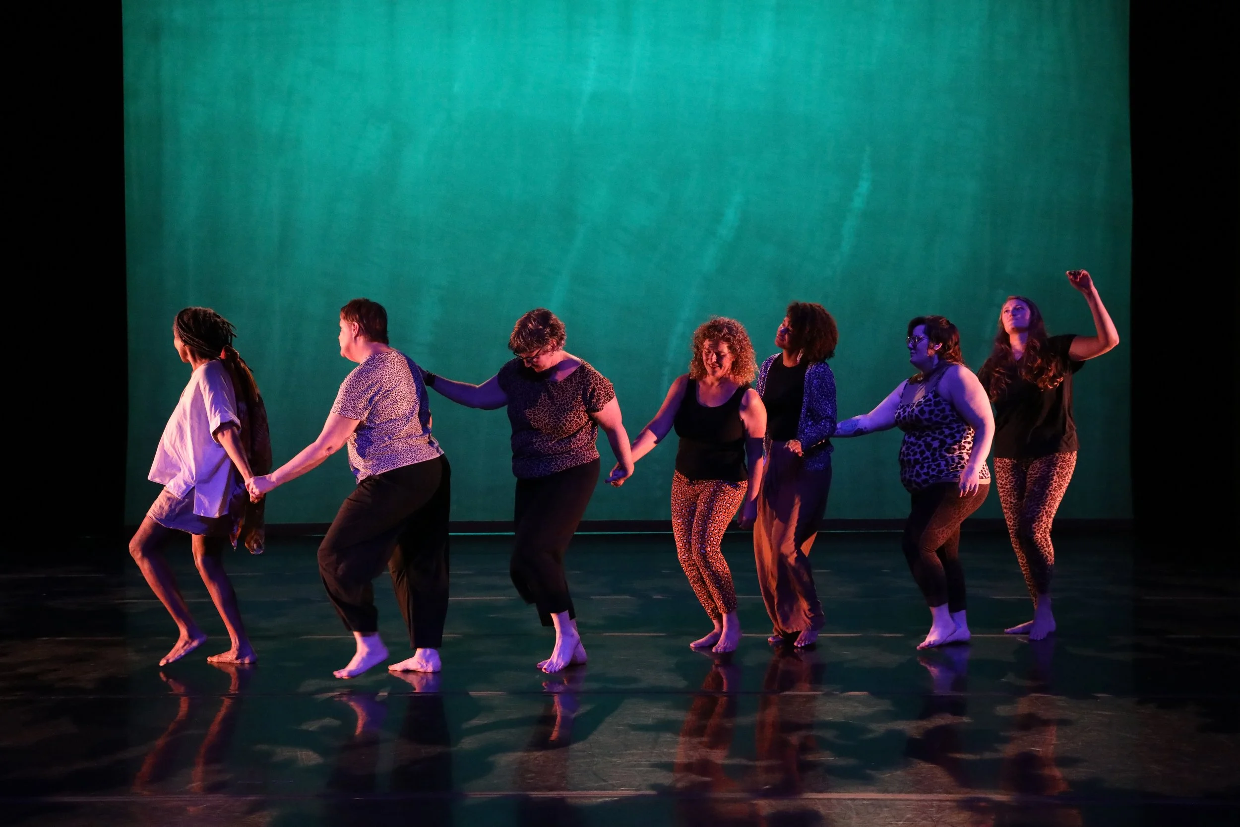 A multiracial group of women in a line holding hands or shoulders dancing across a stage with a turquoise background.