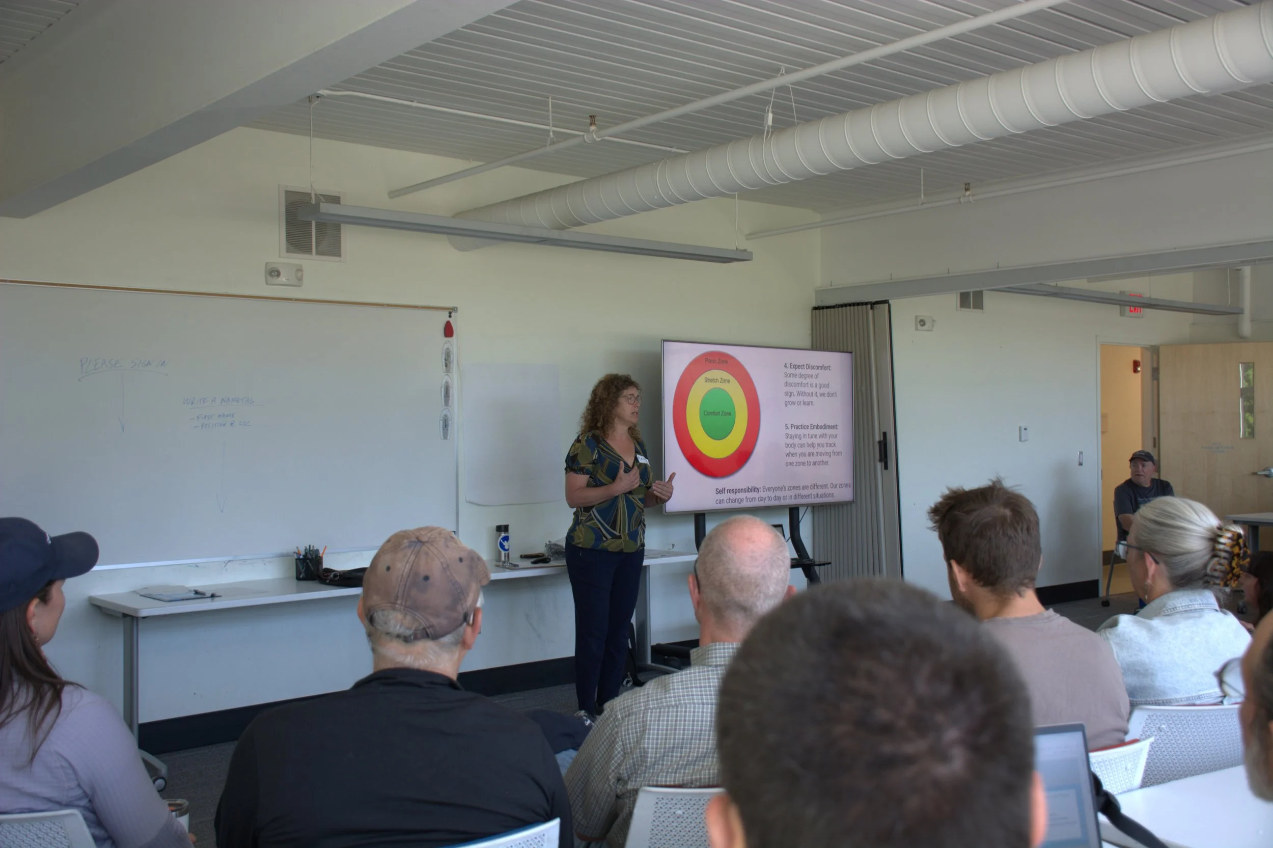 A white woman with curly brown hair and glasses stands in front of a group of people. Next to her is a slide of a bulls eye and text that is too small to read.