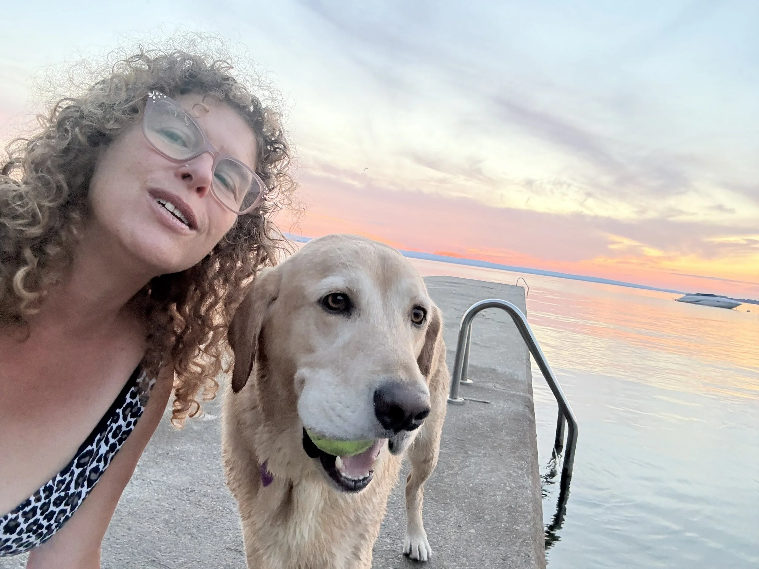 Rachel and her dog on a concrete dock with a lake and sunset in the background. The dog has a ball in his mouth.