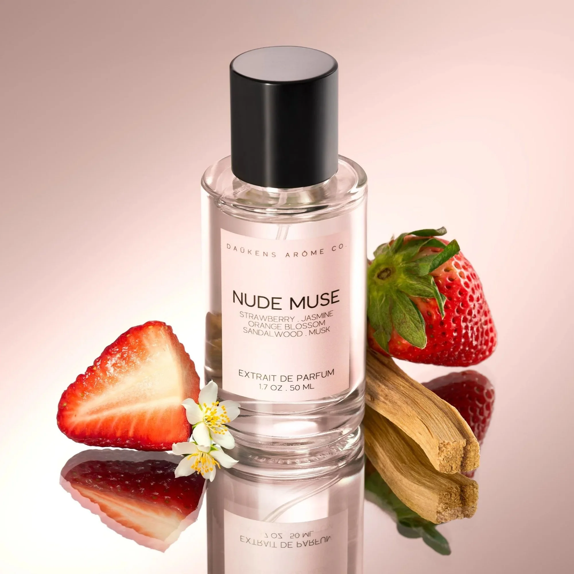 A bottle of perfume labeled 'Nude Muse' surrounded by strawberries, a piece of cinnamon, and white flowers on a reflective surface.