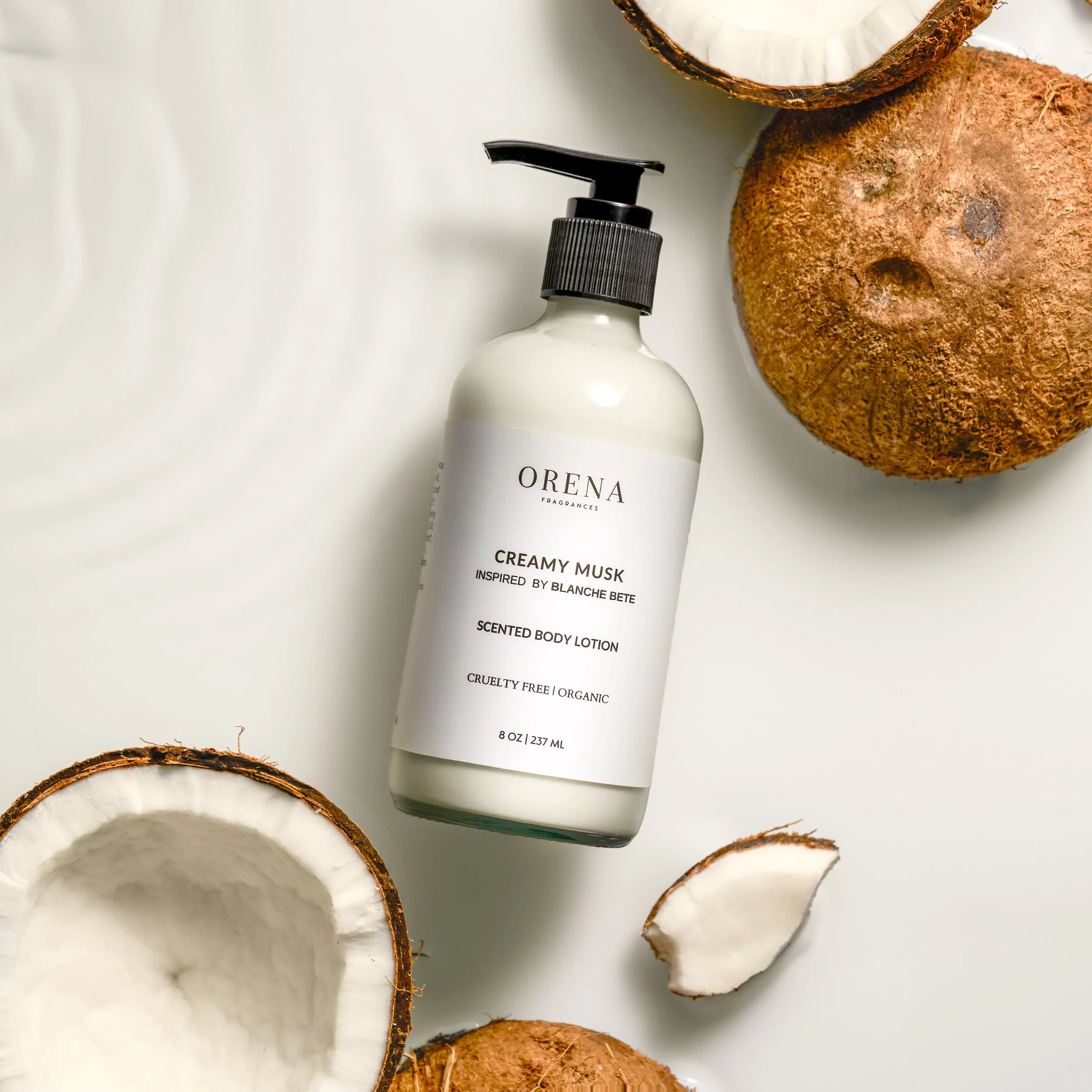 A bottle of Orena Creamy Musk scented body lotion surrounded by whole and halved coconuts on a white background.